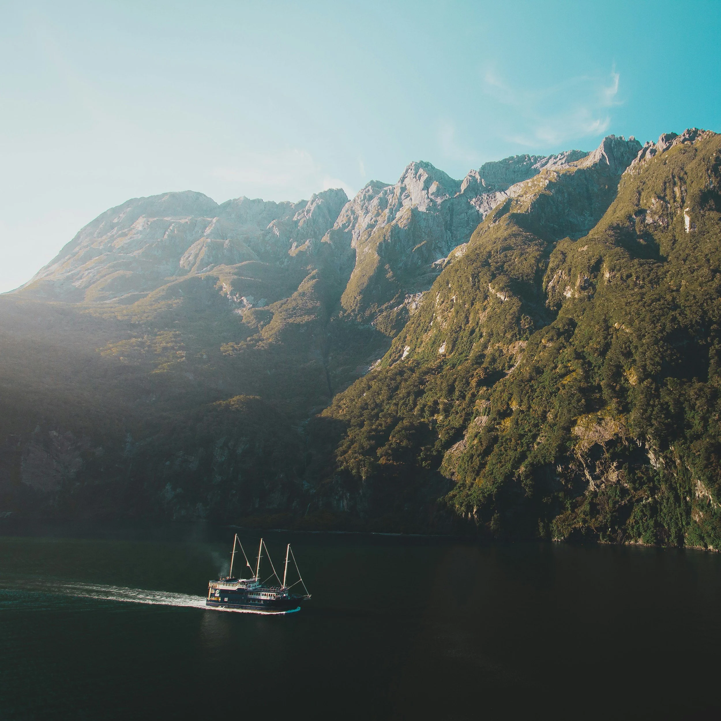 A boat sailing on dark water with a lush green mountain and rocky cliffs in the background under a blue sky with wispy clouds.