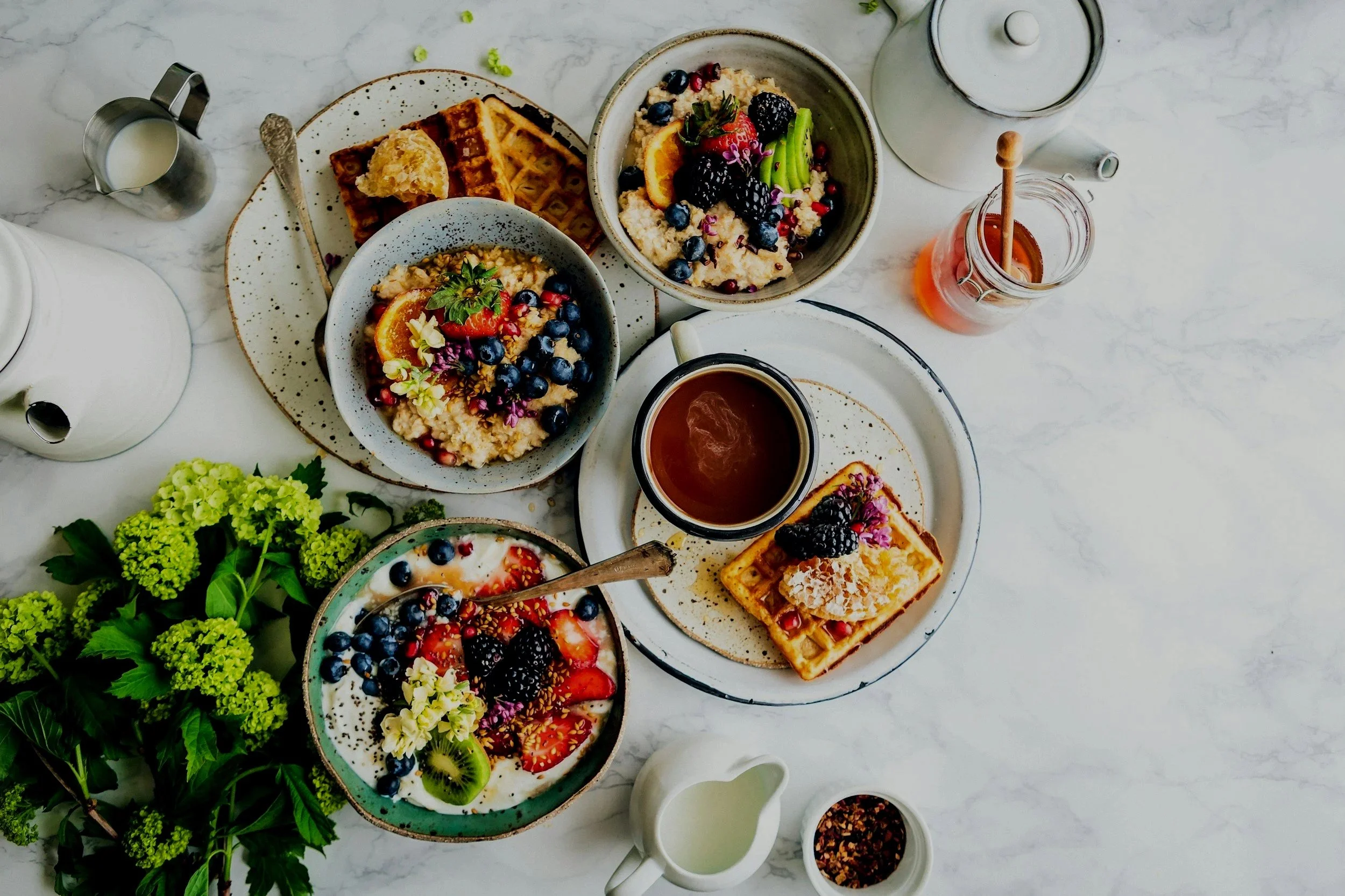 Assorted breakfast items including waffles with toppings, oatmeal with berries and fruit, fresh fruit slices, and various beverages, all on a white marble surface with green floral decoration.