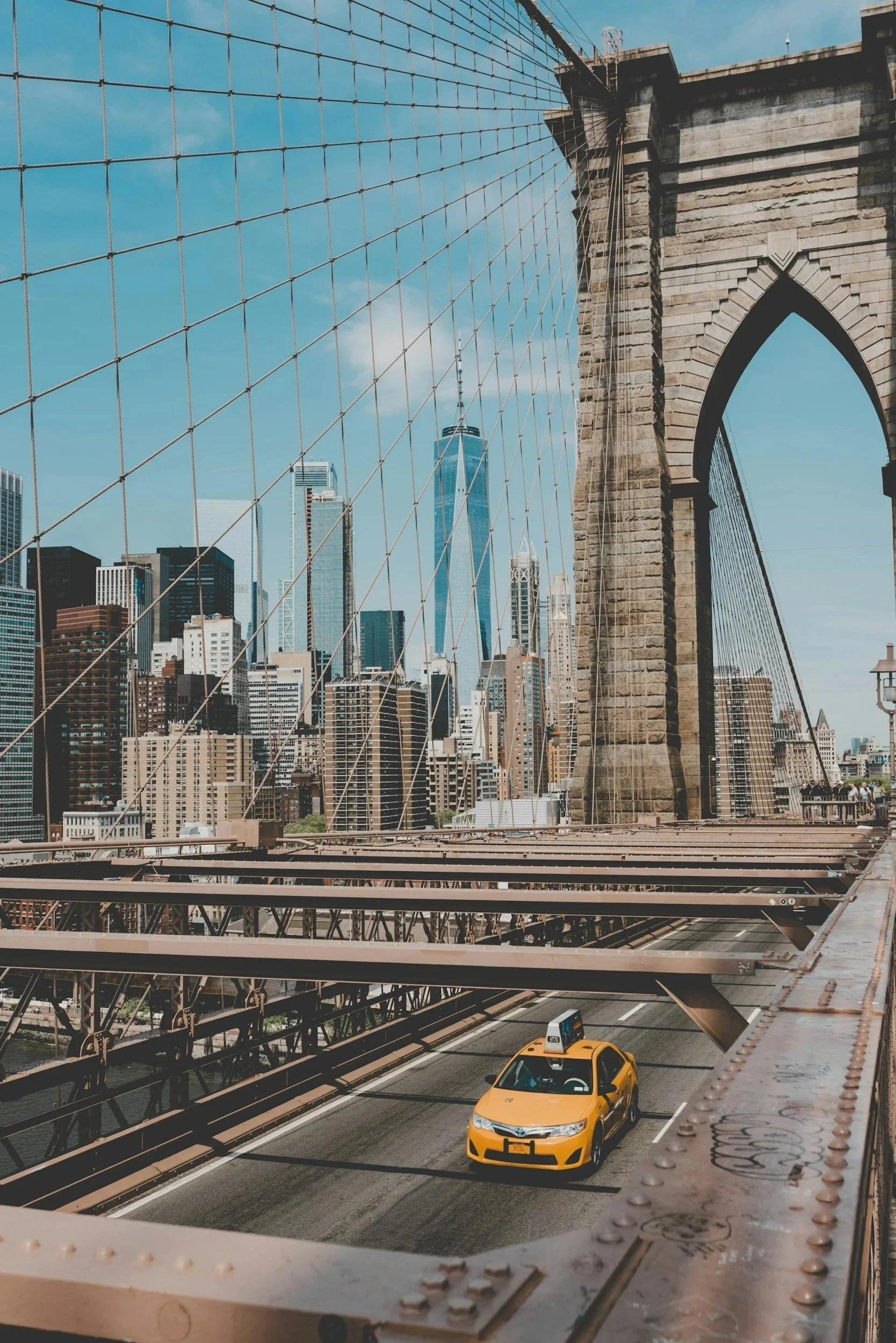 View from a bridge showing a city skyline with tall buildings, one of which is the One World Trade Center, under a partly cloudy blue sky. The bridge has a stone arch and metal suspension cables.