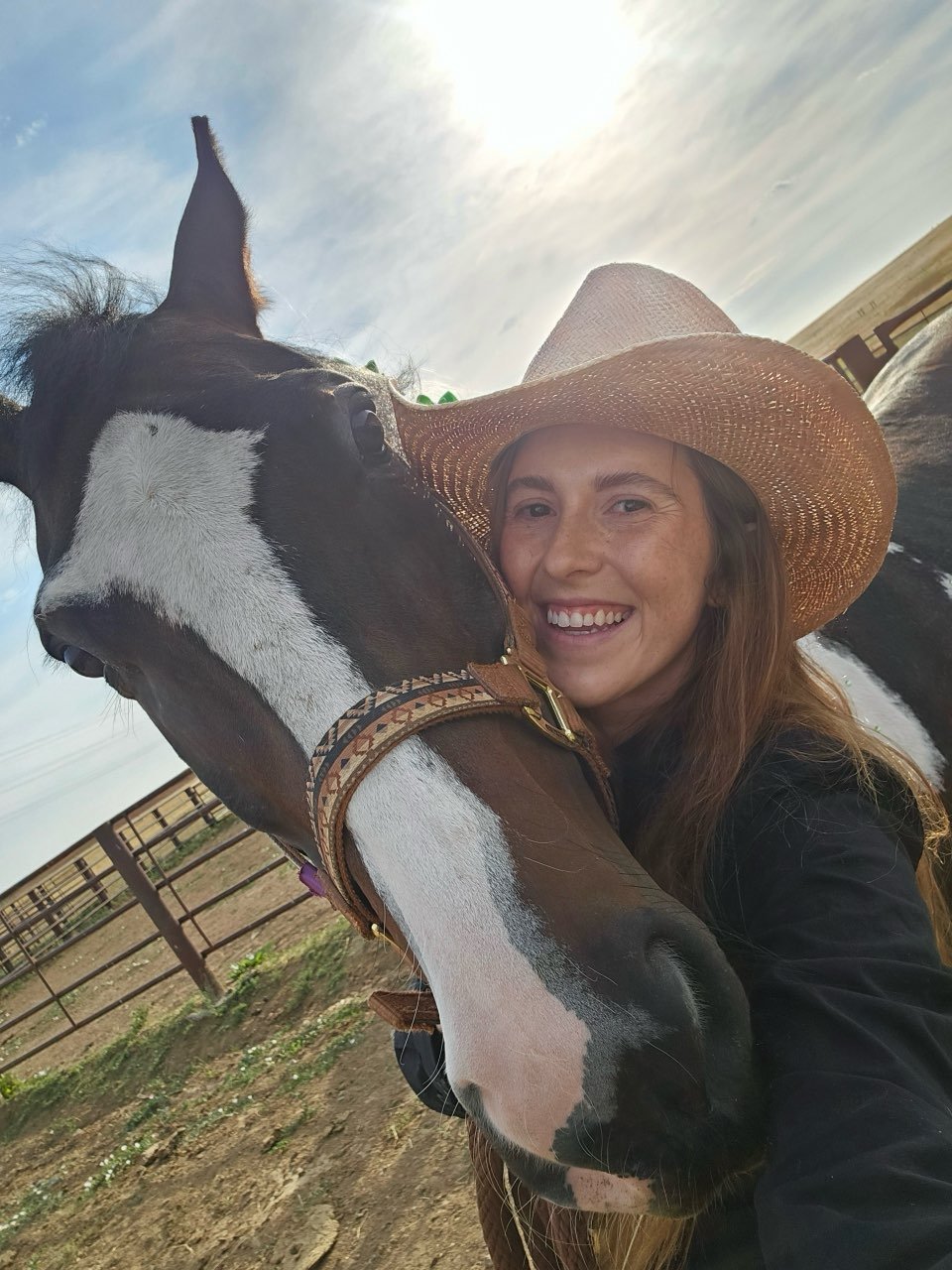 A woman wearing a wide-brimmed straw hat smiling and taking a selfie with a large horse on a farm, with a fence and open land in the background under a partly cloudy sky.
