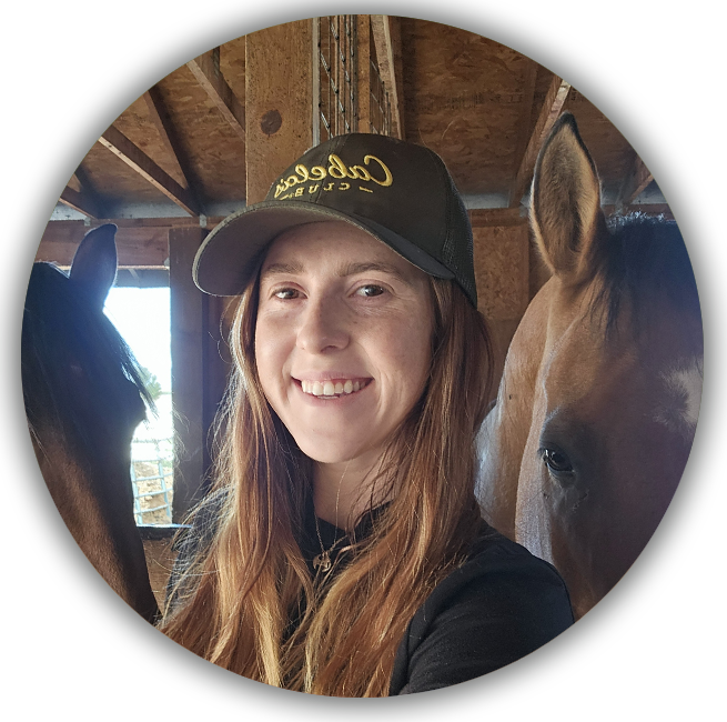 A young woman with long red hair wearing a black cap and shirt, smiling, standing between two horses inside a wooden barn.