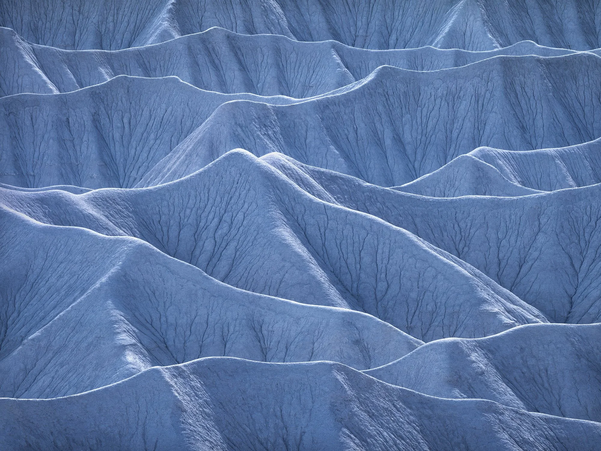 Blue-toned desert badlands with layered ridges and intricate erosion lines forming soft, repeating wave patterns.