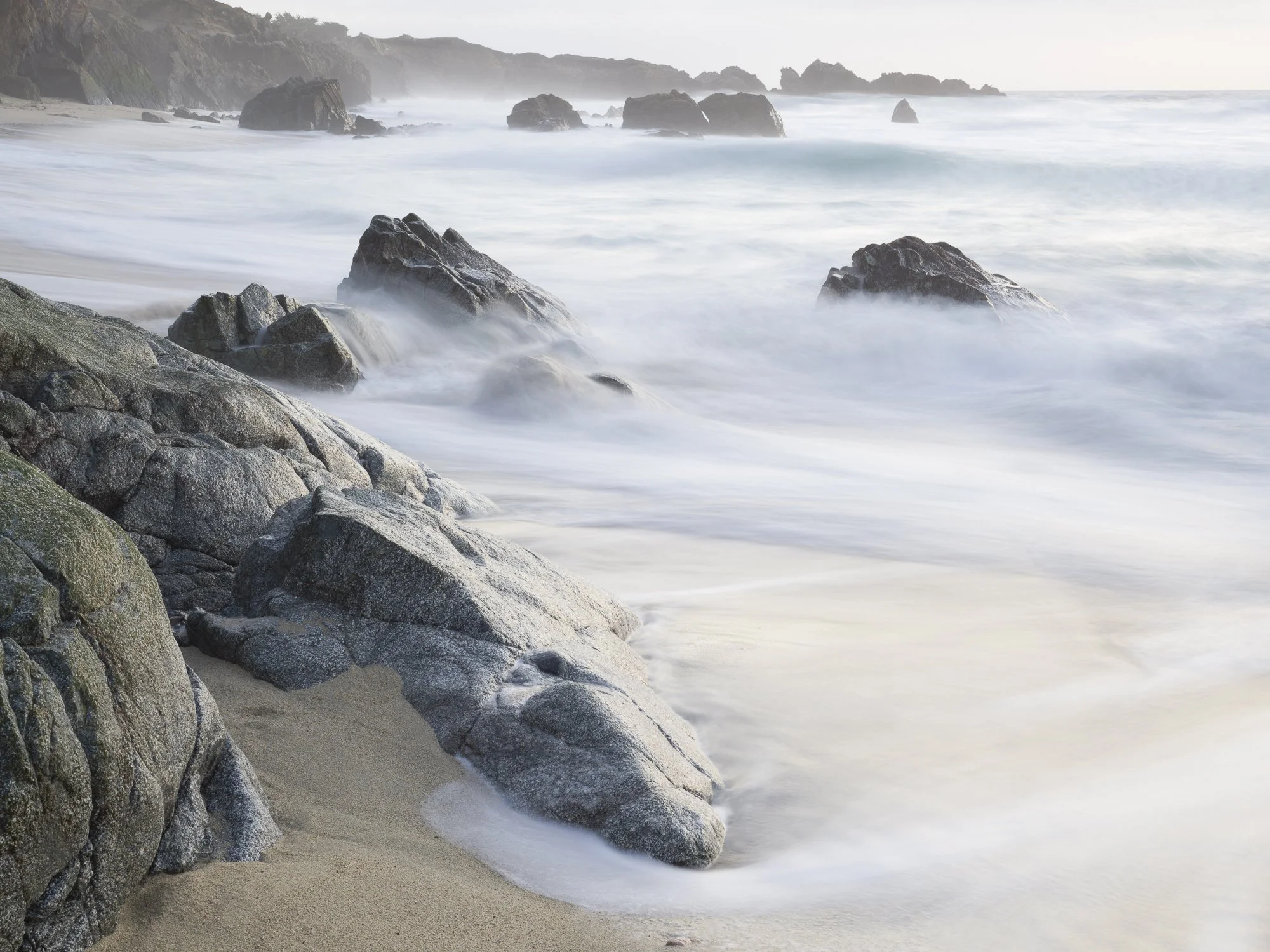 Beach with large rocks, waves washing over the sand, and a foggy, distant shoreline.