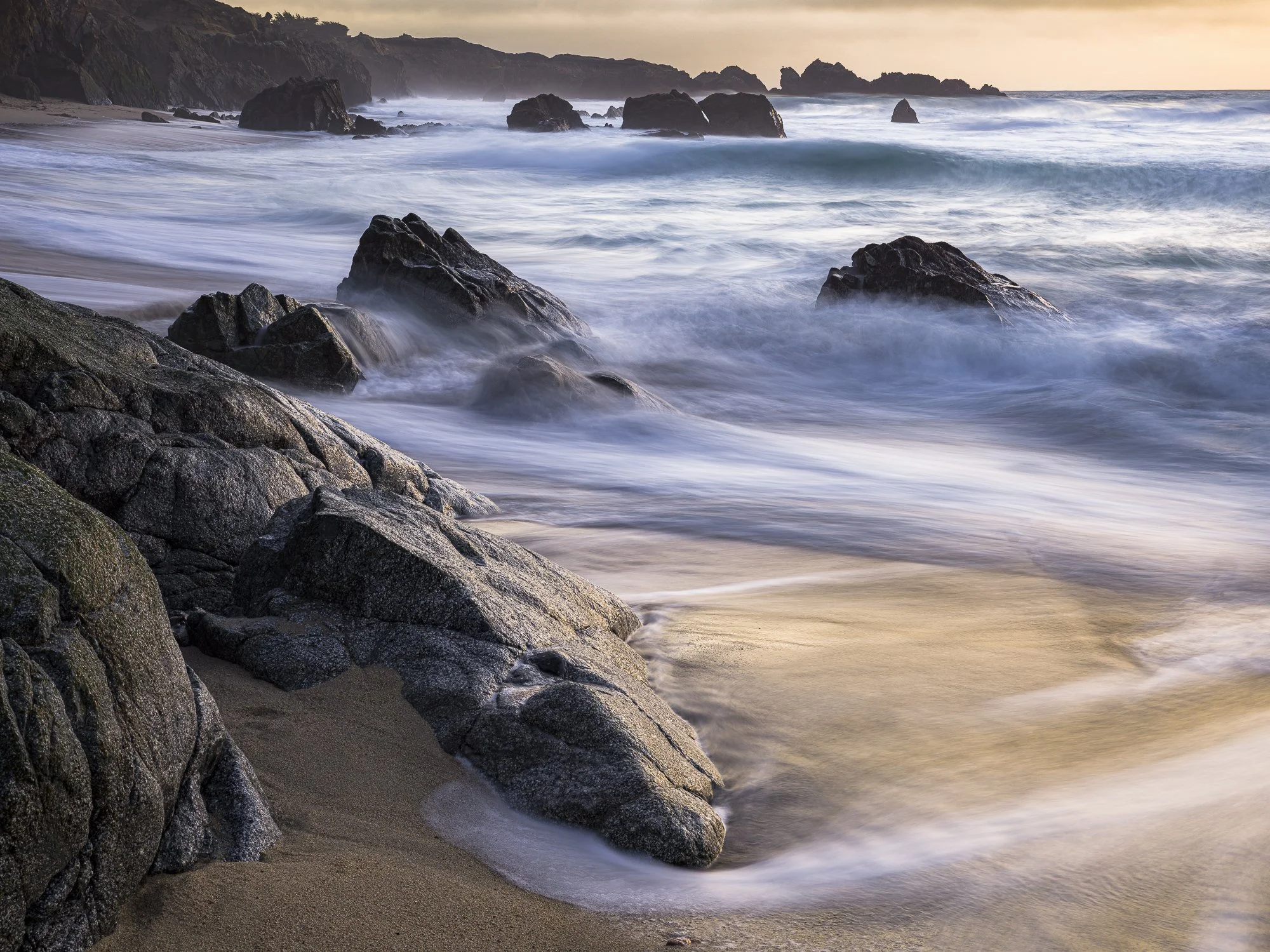 Foamy ocean waves crash against large rocks on a sandy beach at sunset.