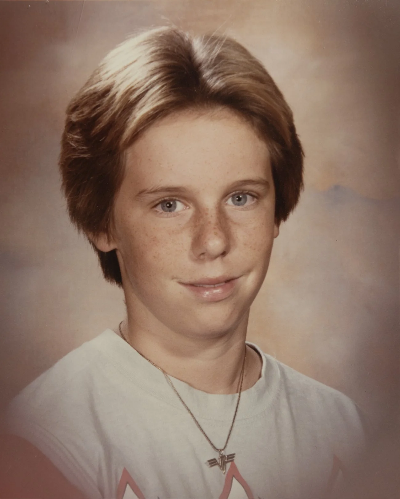 A young boy with short, light brown hair, freckles, and blue eyes, wearing a white t-shirt and a necklace with a pendant, posing against a blurry, warm-toned background.