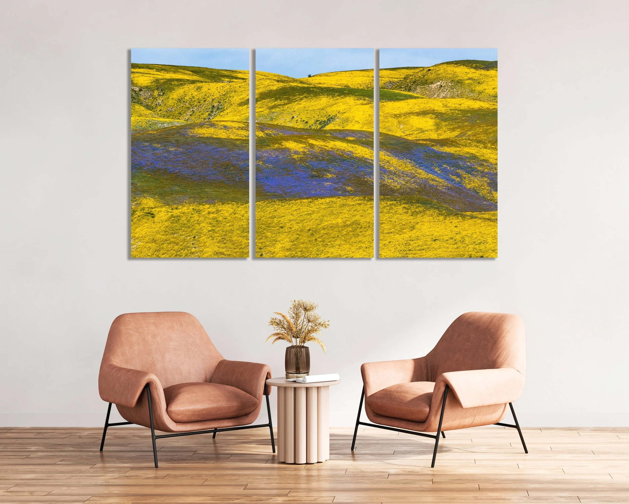 Living room with two pink armchairs, a small round side table with a vase of dried flowers, and a triptych landscape painting of rolling hills with yellow and purple wildflowers on the wall.