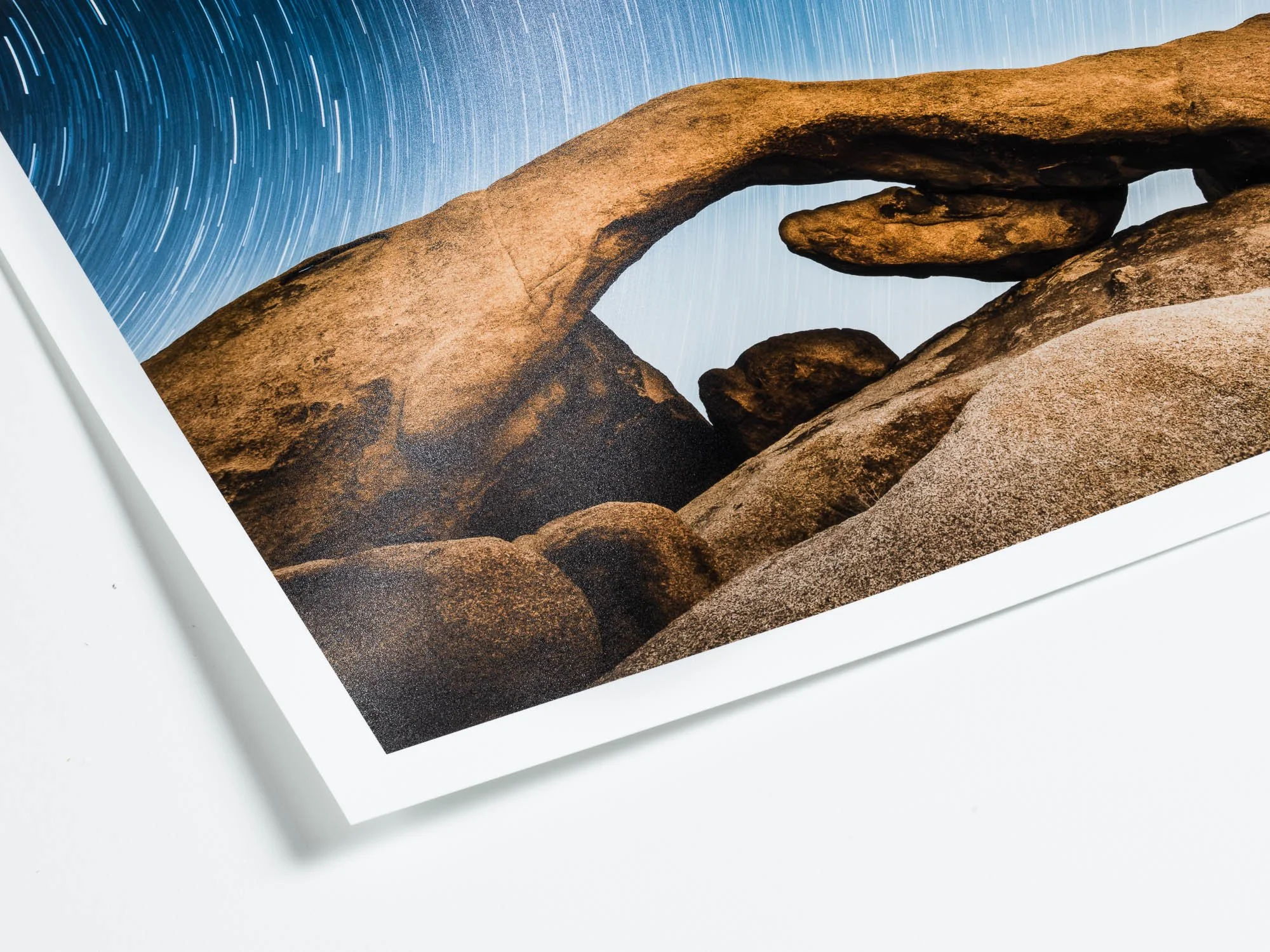 A photograph of a natural rock formation featuring a large arch with a rock balanced underneath it, set against a backdrop of star trails in the night sky.