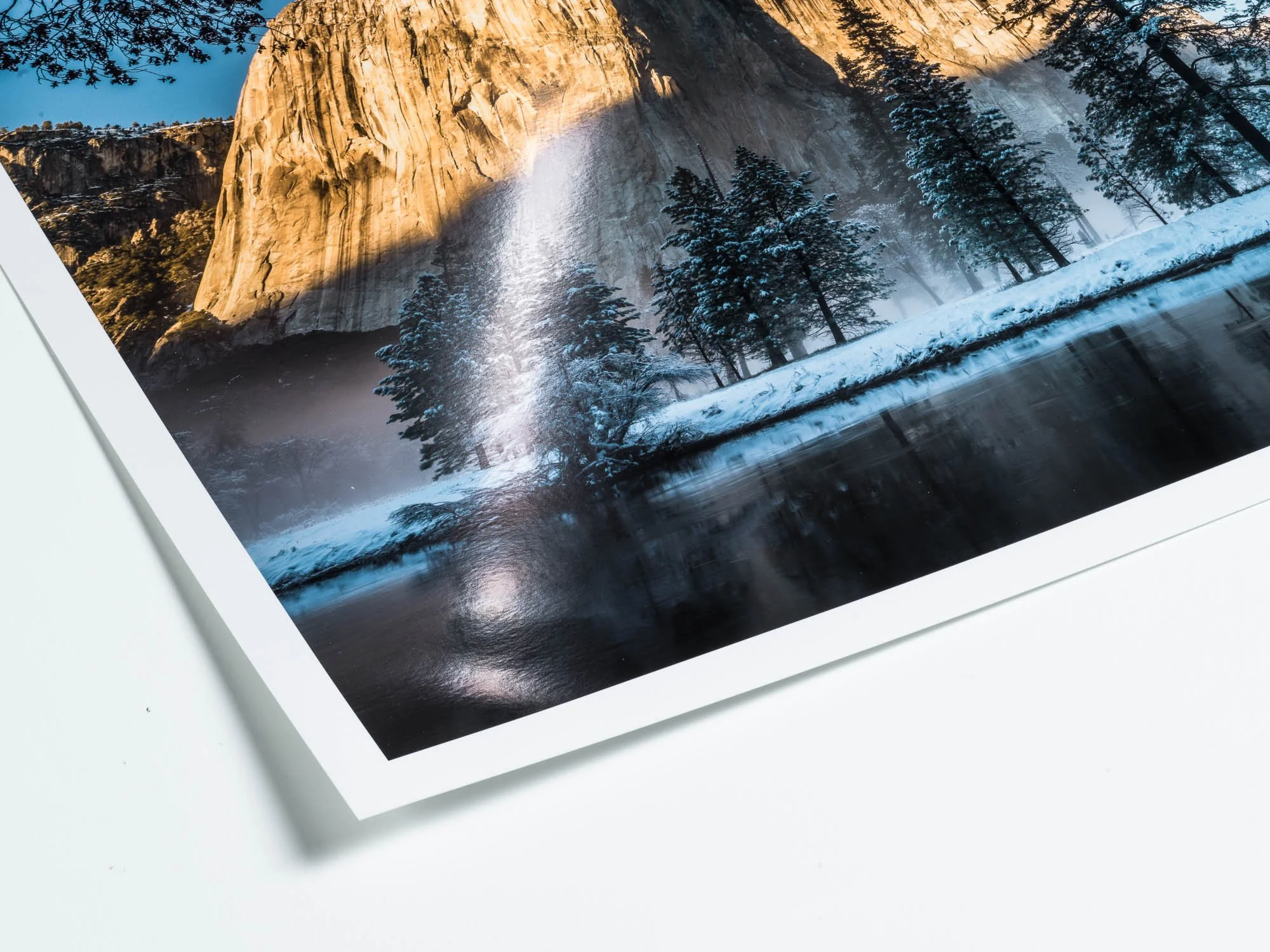 A printed photograph of a snowy landscape with trees, a waterfall, and a large rock face, reflected in a body of water.