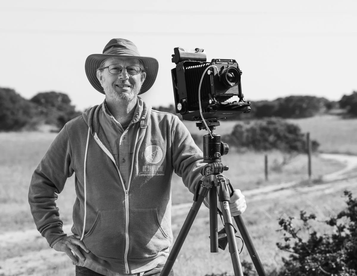 Portrait of Kevin Osborn, photo instructor, standing outdoors with a camera mounted on a tripod in a black and white photograph.