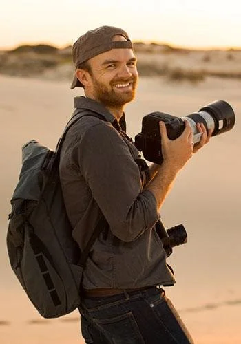 Portrait of photographer Austin Mann carrying a camera and backpack while standing outdoors in a sandy, coastal environment.