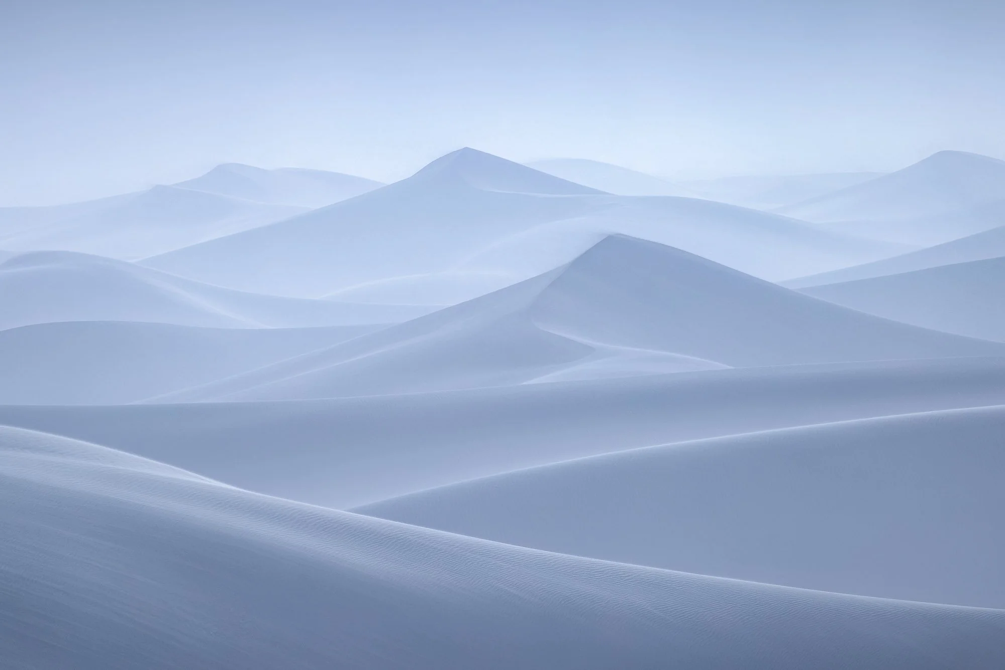 Soft blue sand dunes layered in mist, creating smooth flowing curves in a minimalist desert landscape.