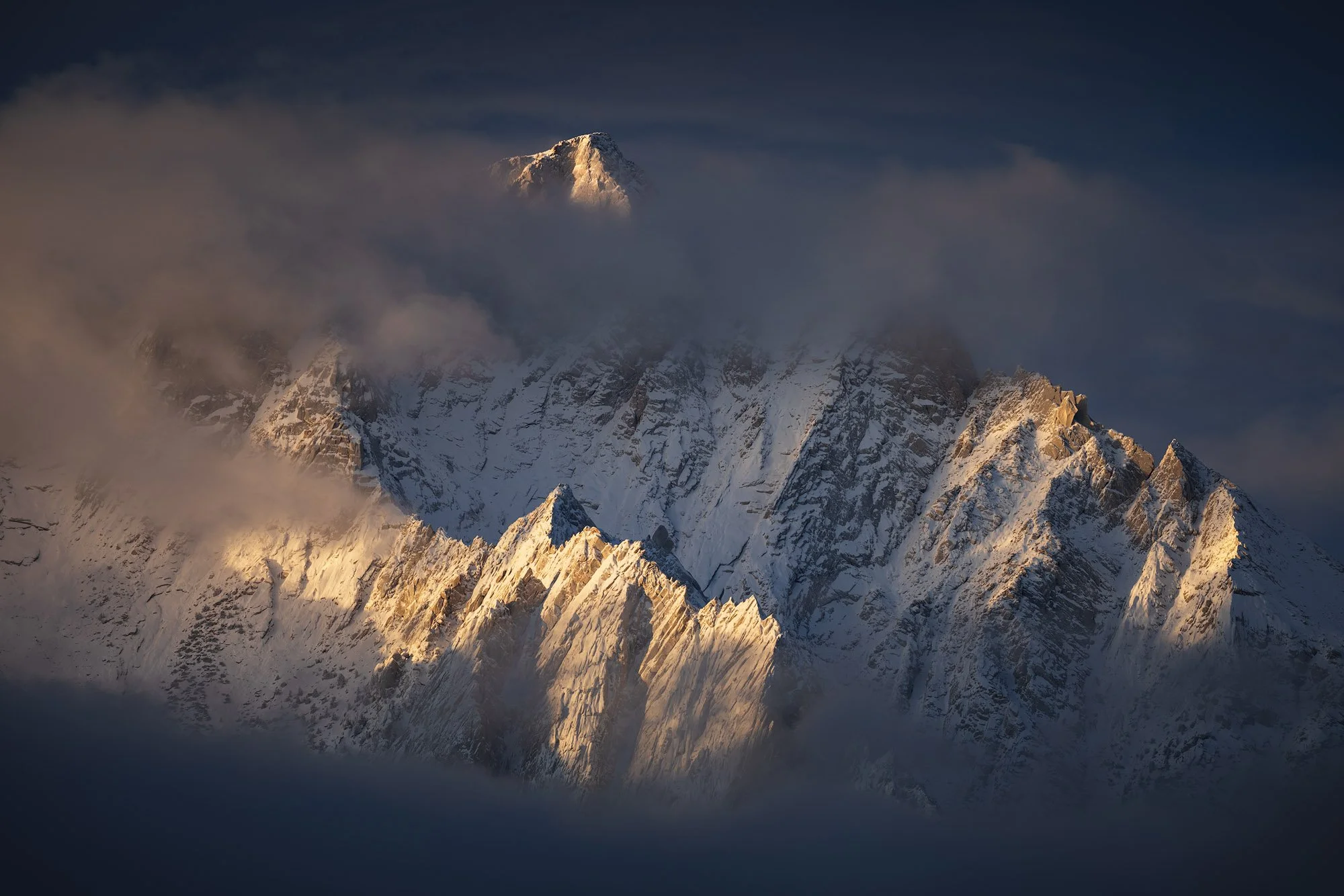 Snow-covered mountain peak emerging through clouds, with golden sunrise light illuminating sharp alpine ridges.