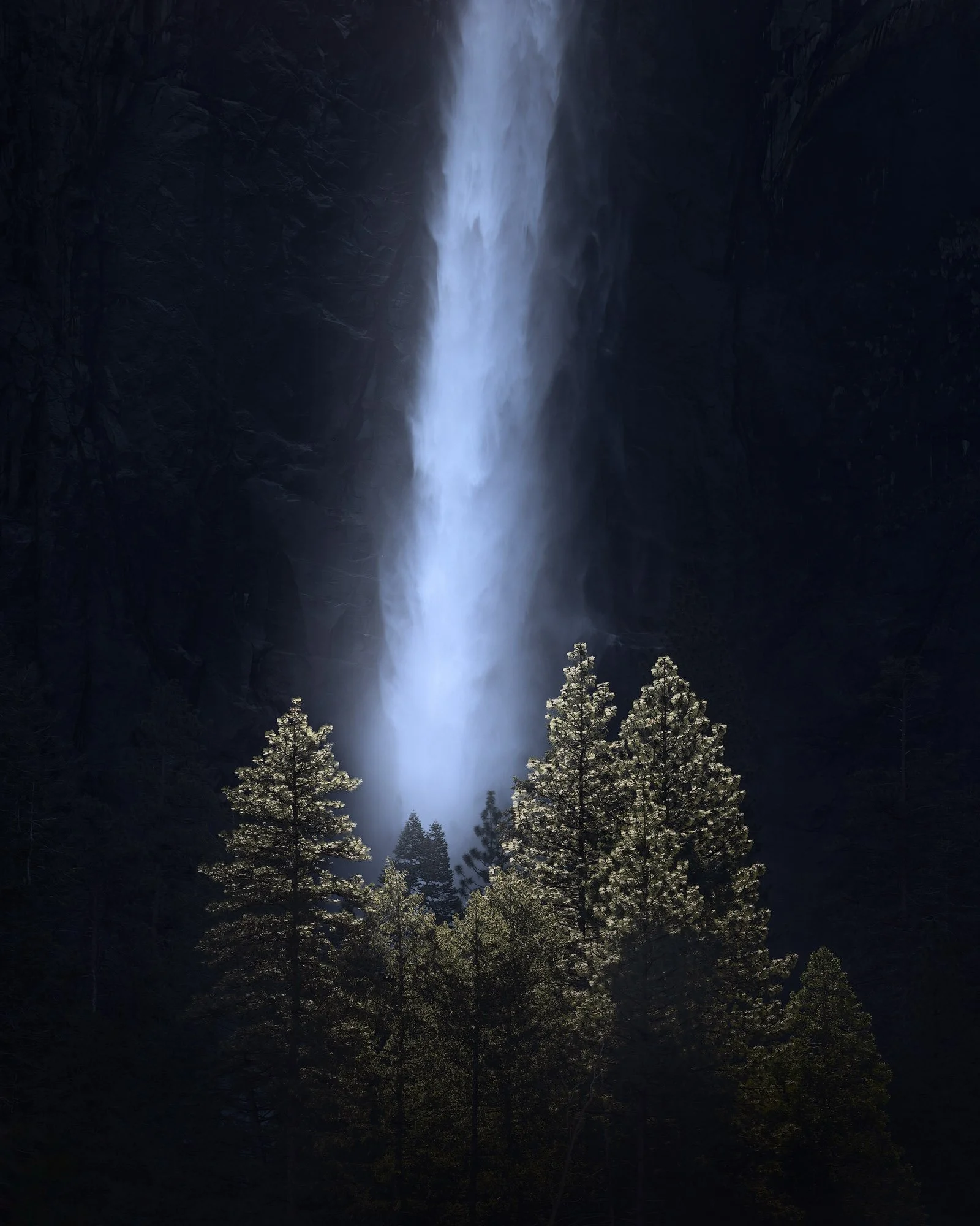 Towering waterfall plunging between dark granite cliffs, illuminating a stand of pine trees below in dramatic, moody light.