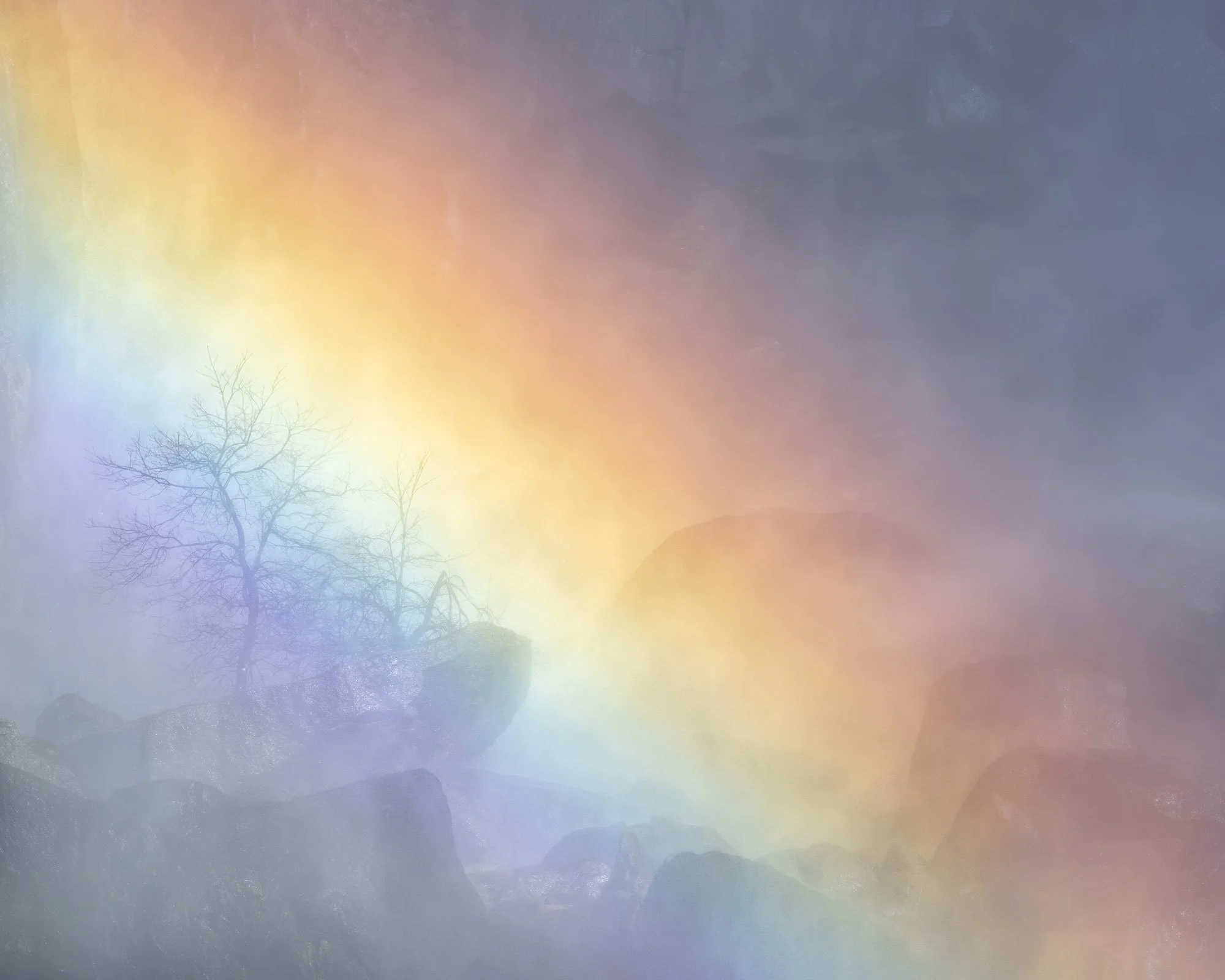 A soft rainbow arcs through mist over silhouetted trees and rocks, creating a dreamy, pastel landscape.