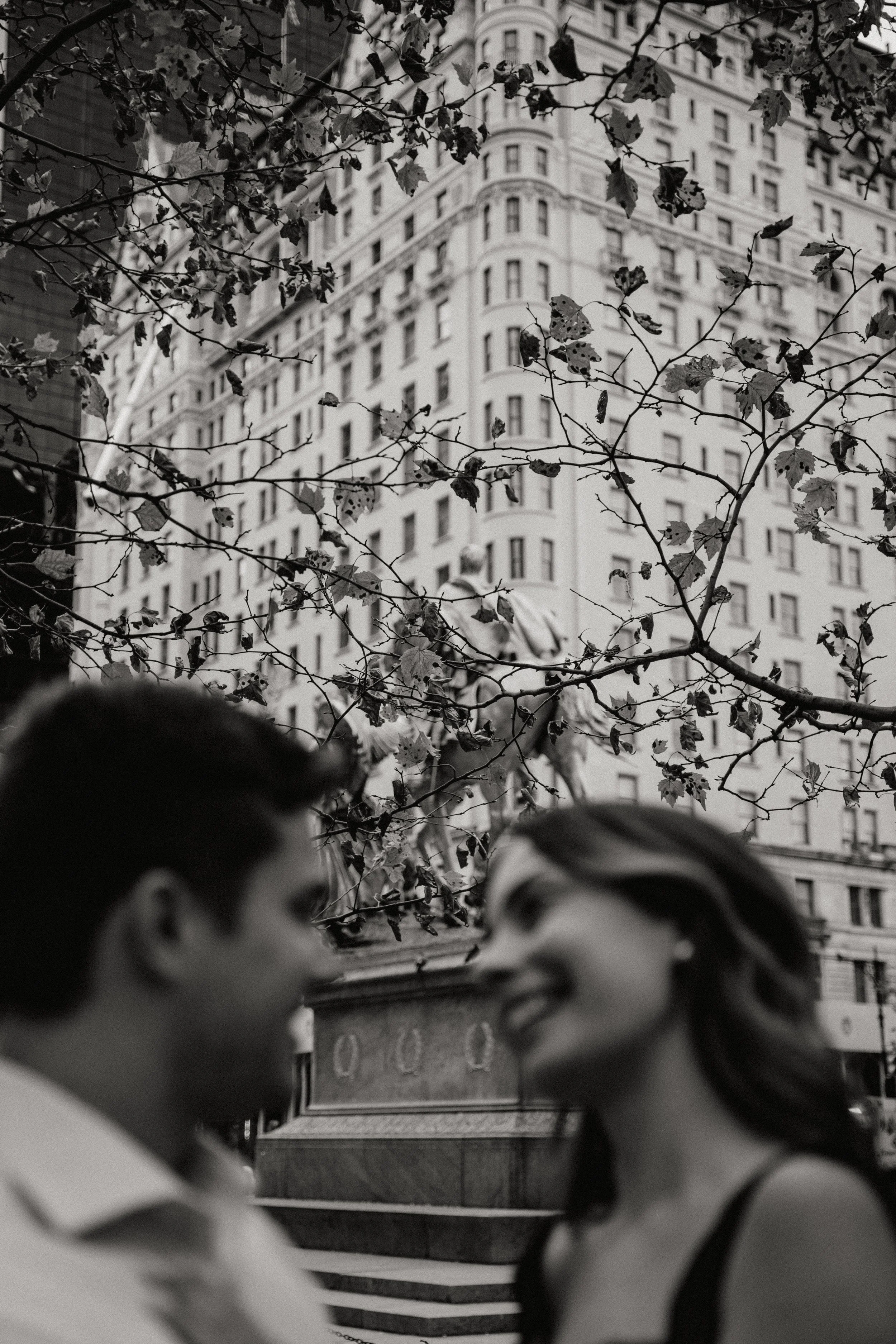 Black and white photo of a young couple smiling and looking at each other, with a city building and tree branches in the background in Somerville, New Jersey photographed by Dominique Paulette Photography