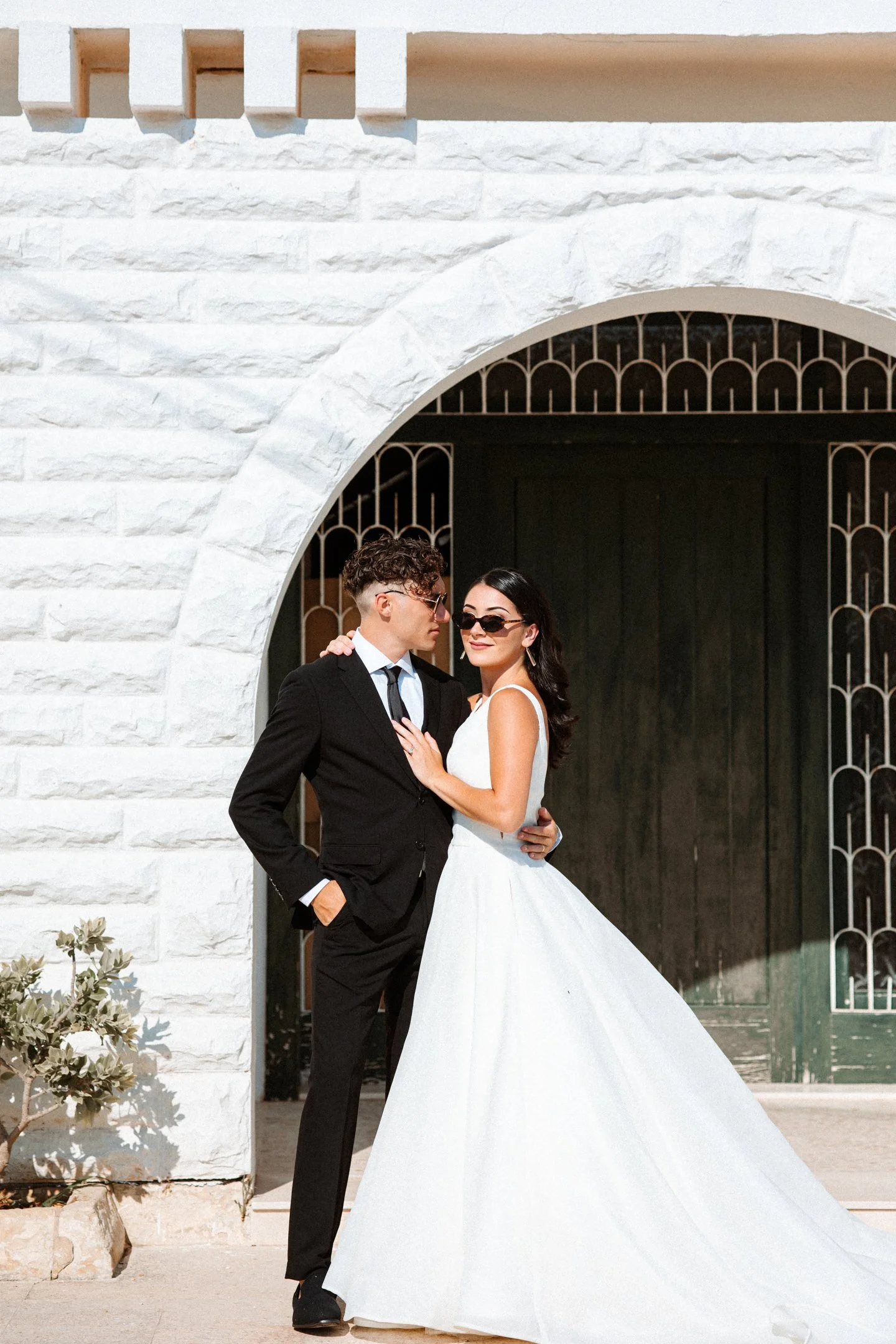 A man and woman in wedding attire standing close together in front of a building with a white brick archway and black wrought iron gate at their wedding in Malta, photographed by Dominique Paulette Photography