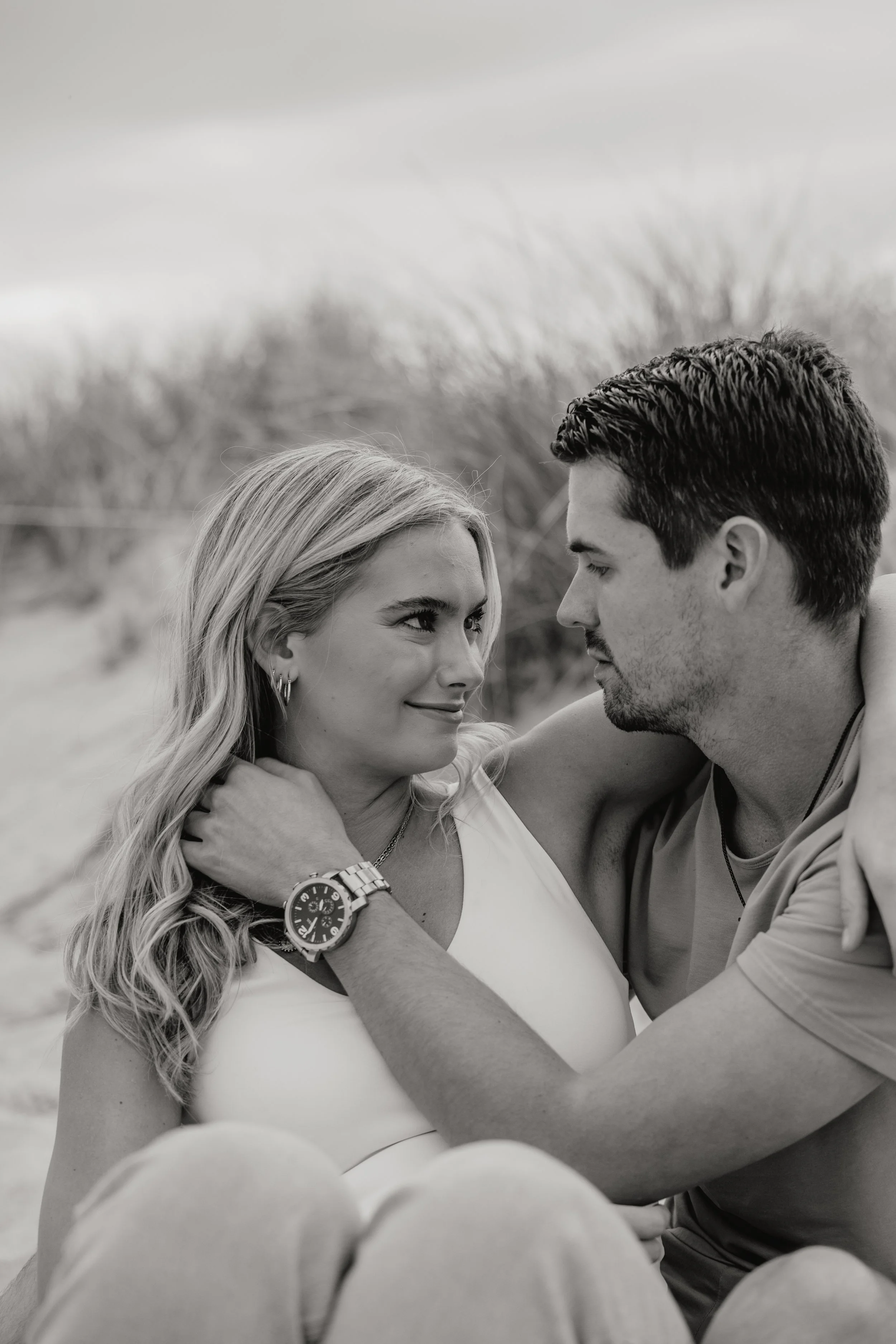 A black and white photo of a couple sitting close together outdoors, looking at each other lovingly, on a beach with grass in the background for their engagement photos by the Jersey Shore photographed by Dominique Paulette Photography