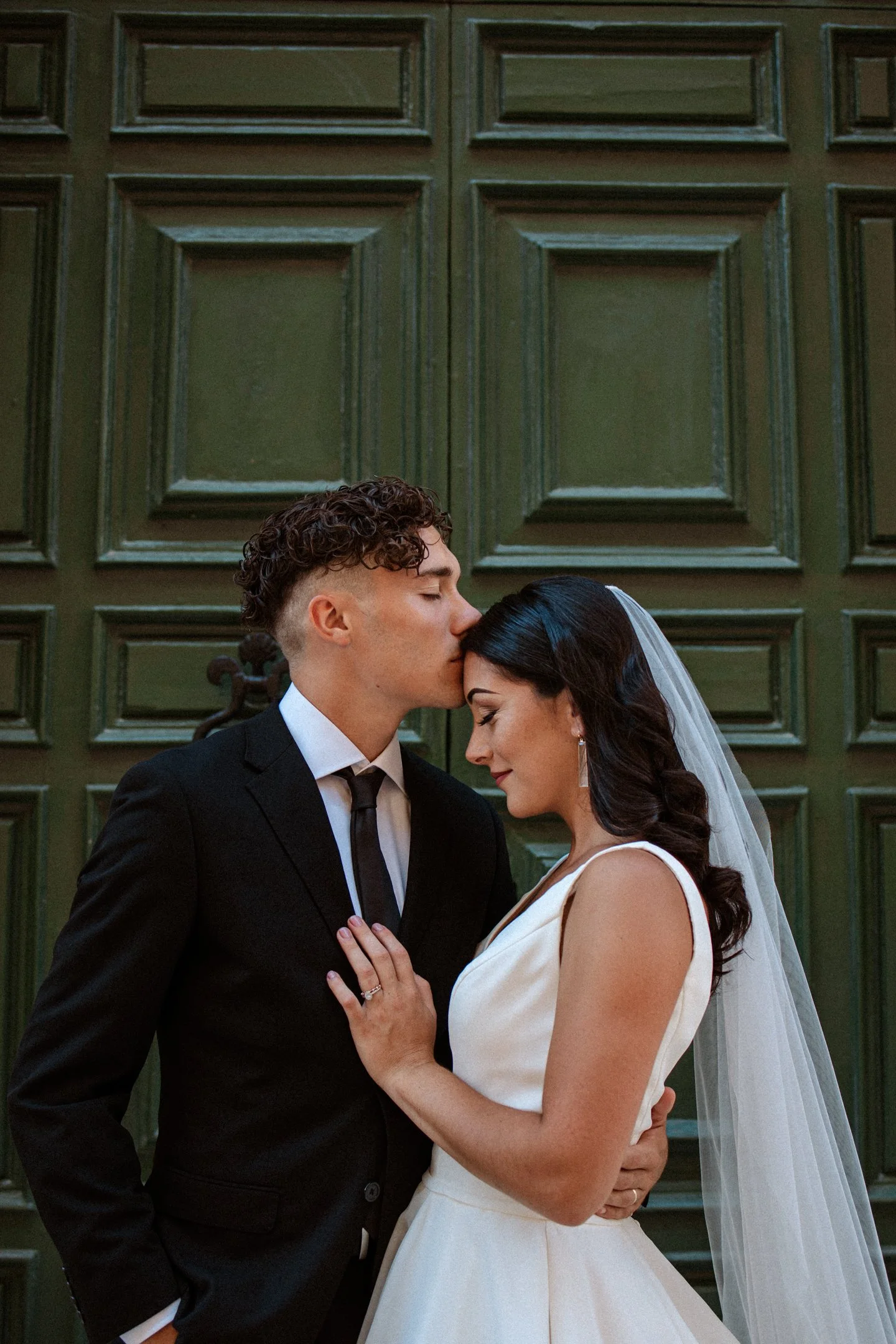 A newlywed couple in wedding attire standing closely together in front of a green paneled door, with the groom gently kissing the bride's forehead for their wedding in Malta, photographed by Dominique Paulette Photography