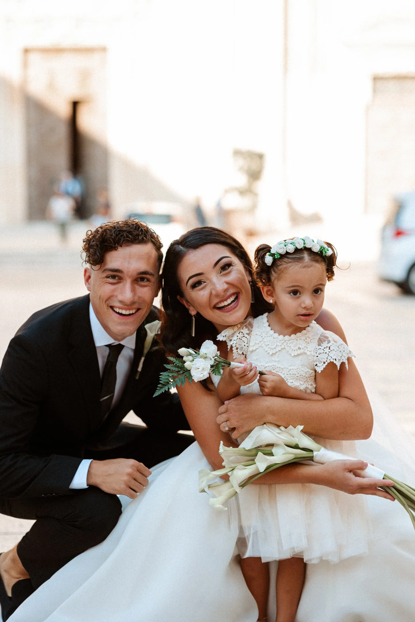 A happy bride, groom, and young girl in a wedding outdoors, with the bride holding flowers and the girl wearing a lace dress and floral headband at their wedding in Malta, photographed by Dominique Paulette Photography