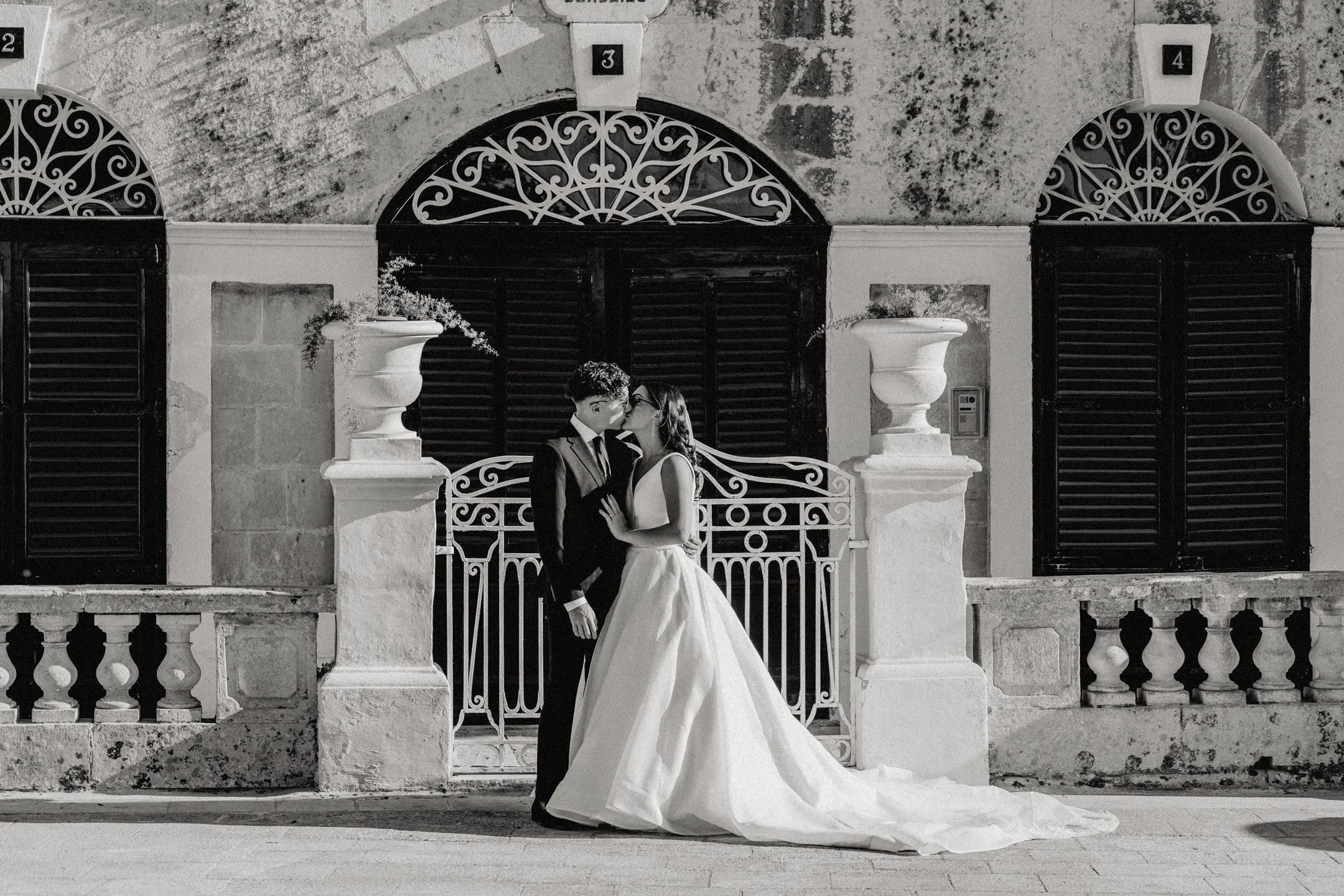 A couple dressed in wedding attire sharing a kiss in front of an ornate iron gate and stone building in black and white at their wedding in Somerville, New Jersey, photographed by Dominique Paulette Photography