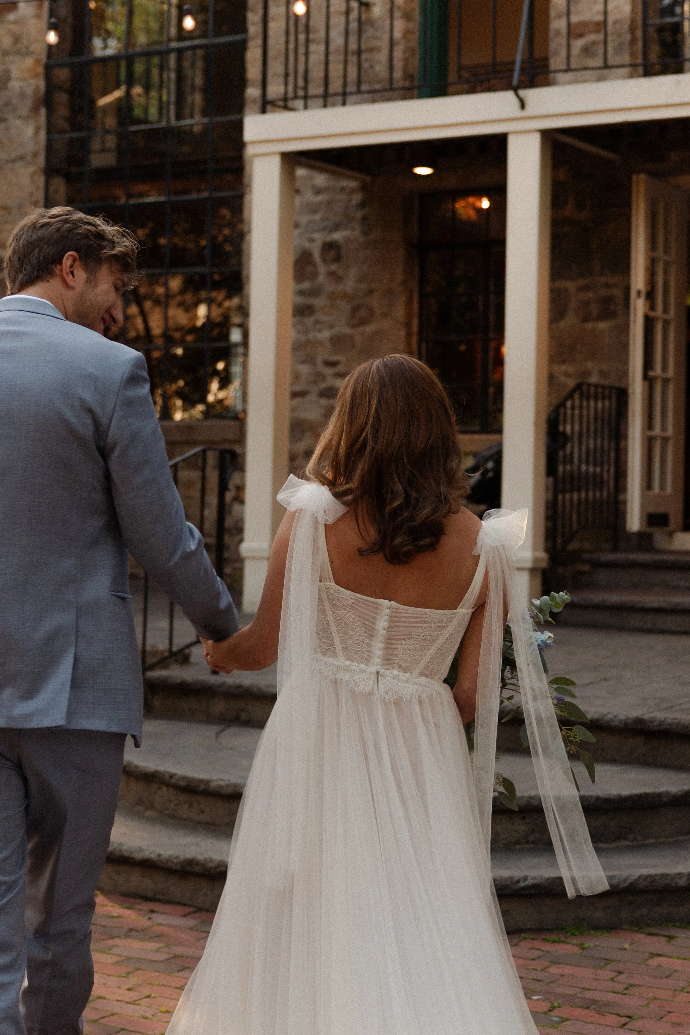 A couple holding hands outside a rustic building at sunset, with the woman wearing a white wedding dress and holding a bouquet at a wedding in Somerville, New Jersey photographed by Dominique Paulette Photography