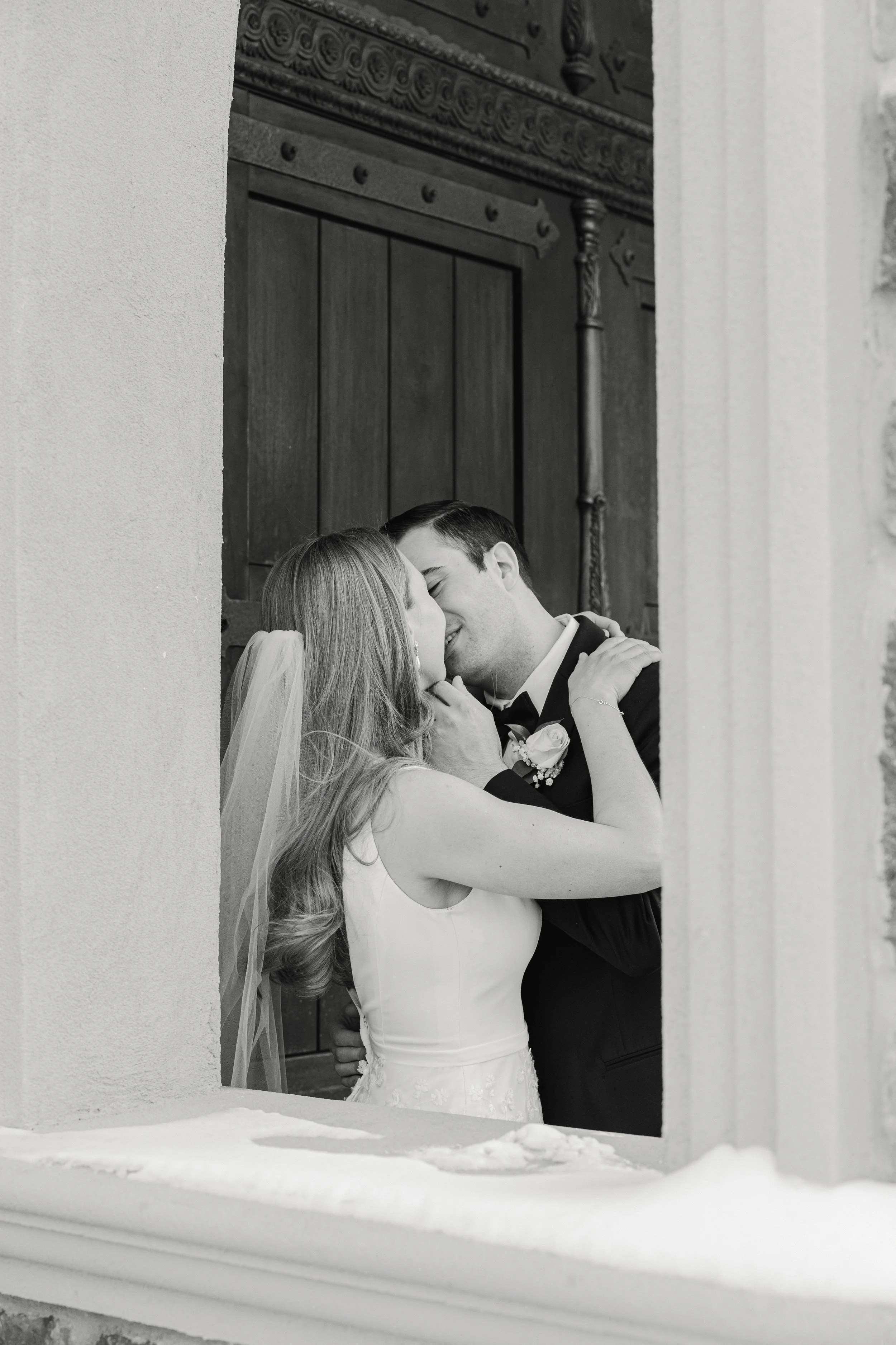 A black and white photo of a bride and groom sharing a kiss indoors, viewed through a window with snow on the sill at their wedding in Somerville, New Jersey, photographed by Dominique Paulette Photography