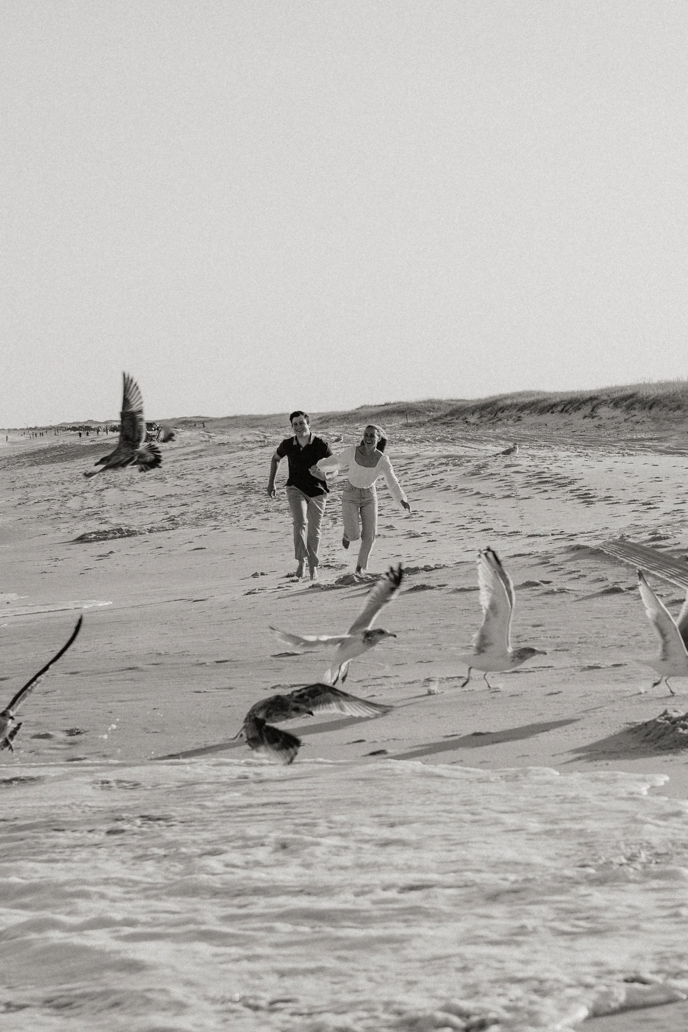 A black-and-white photo of a couple running on a sandy beach with seagulls flying around in the foreground and background for their engagement shoot on the Jersey shore, New Jersey photographed by Dominique Paulette Photography