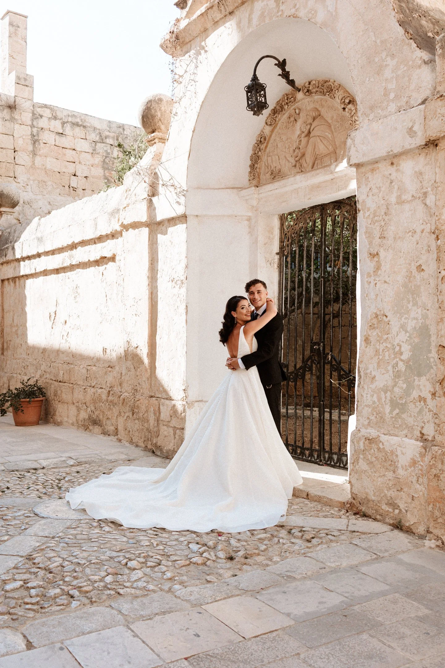 A newlywed couple embraces in front of an old stone building with an arched entrance and metal gate, on a stone-paved street at their wedding in Malta, photographed by Dominique Paulette Photography