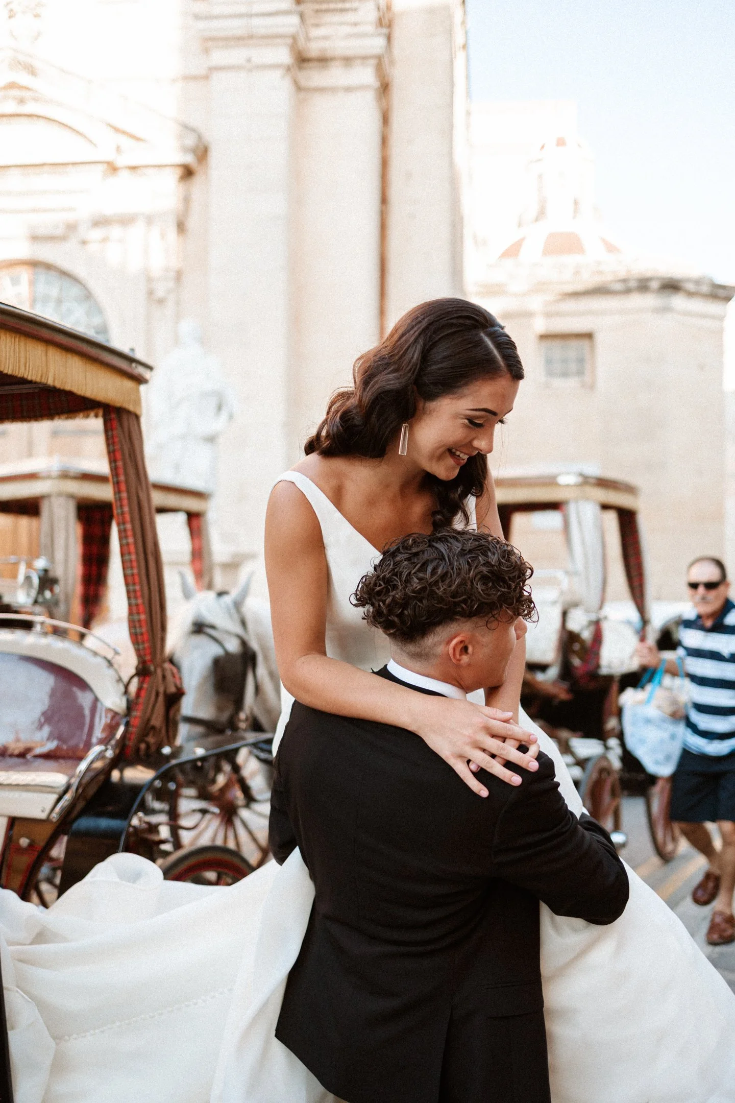 A bride and groom share a dance outdoors with historic buildings and horse-drawn carriages in the background in Malta, photographed by Dominique Paulette Photography