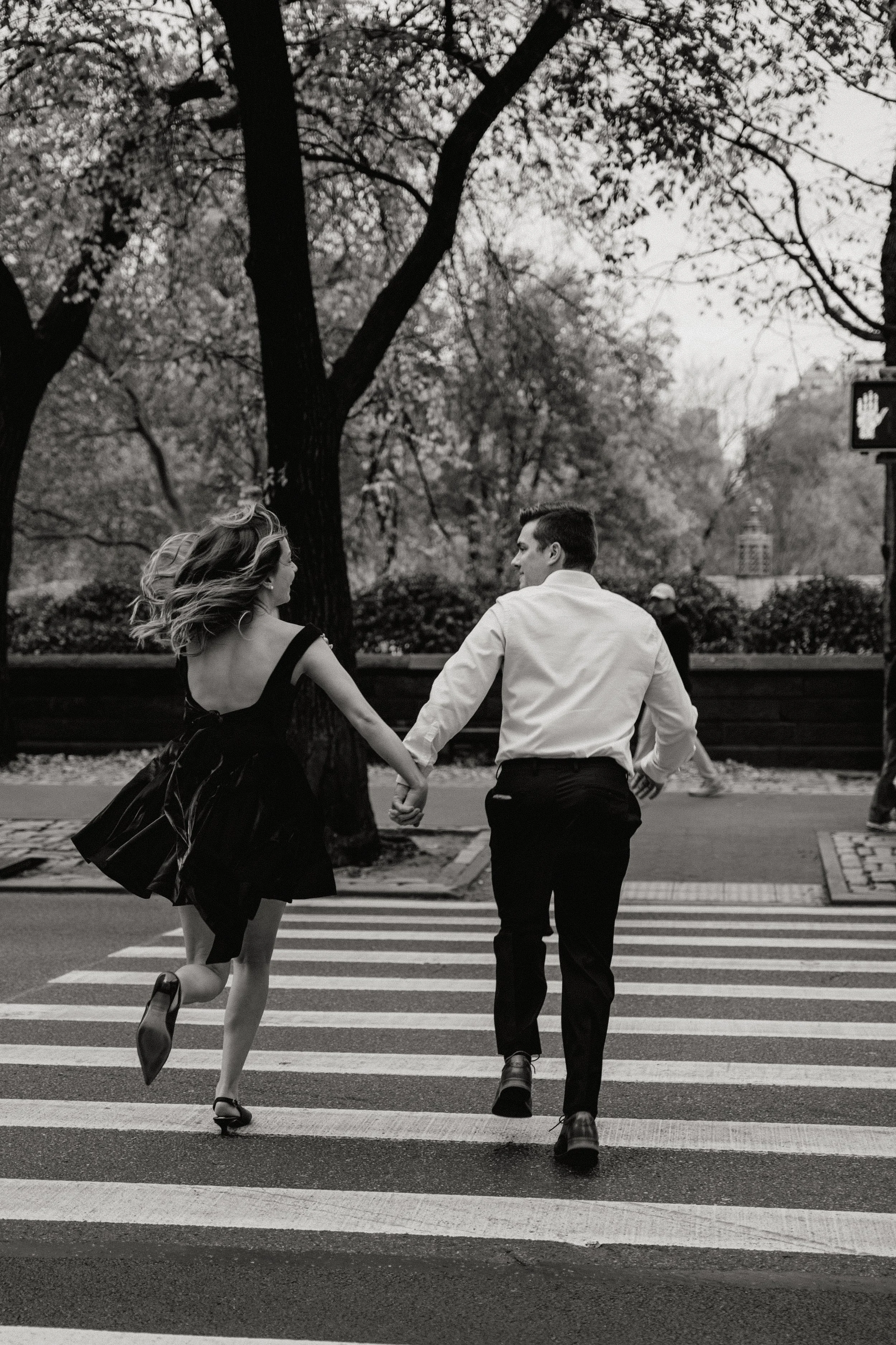 A black and white photo of a couple holding hands while running across a zebra crossing in a park-like setting with trees in the background for their engagement photos photographed by Dominique Paulette Photography