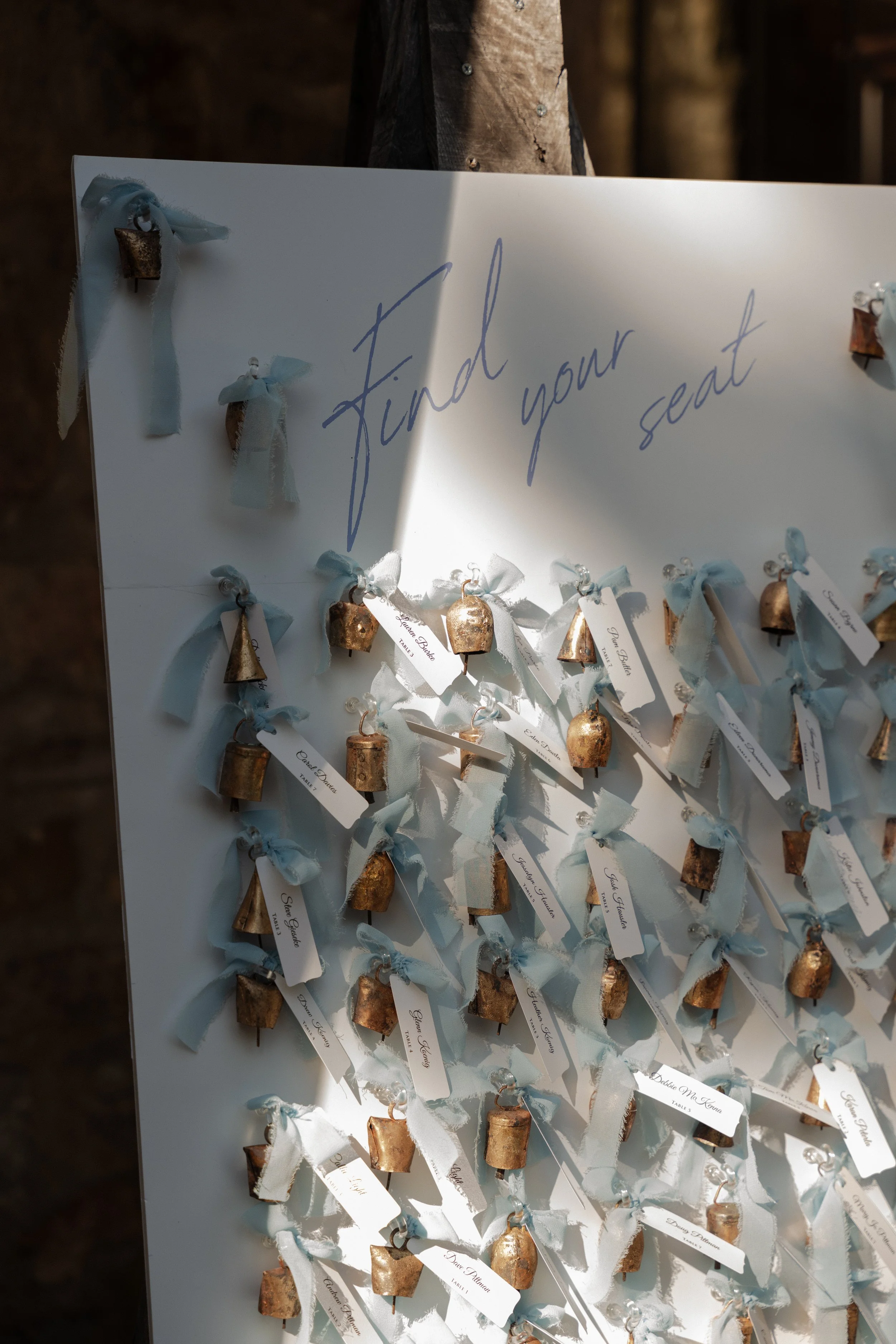 A white board with the phrase 'Find your seat' written at the top, decorated with multiple small bells tied with light blue ribbons at a wedding in Somerville, New Jersey photographed by Dominique Paulette Photography