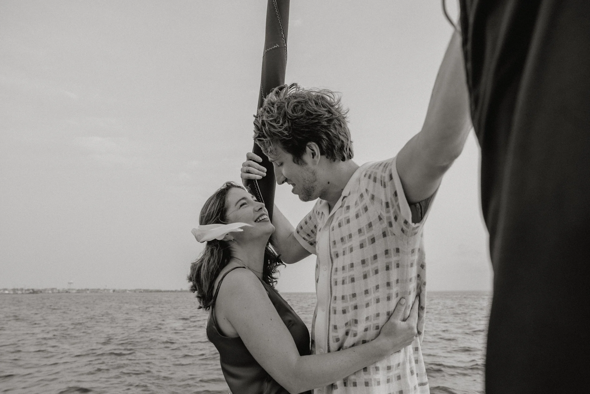 A black-and-white photo of a young couple smiling and looking at each other while embracing by the water, engagement photos in New Jersey photographed by Dominique Paulette Photography
