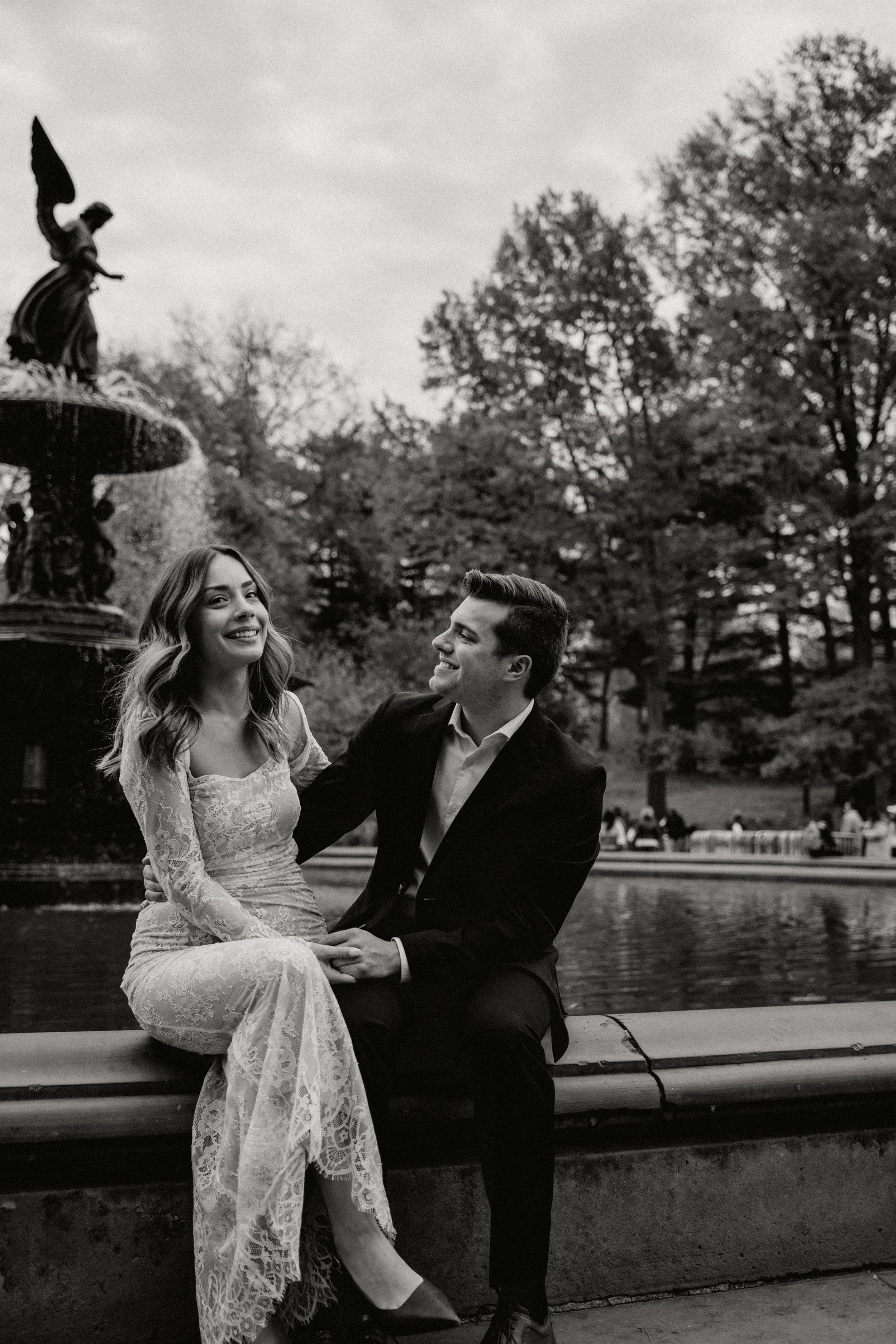 A couple sitting on a park bench near a fountain, sharing a joyful moment, in black and white for their engagement photos in New Jersey photographed by Dominique Paulette Photography