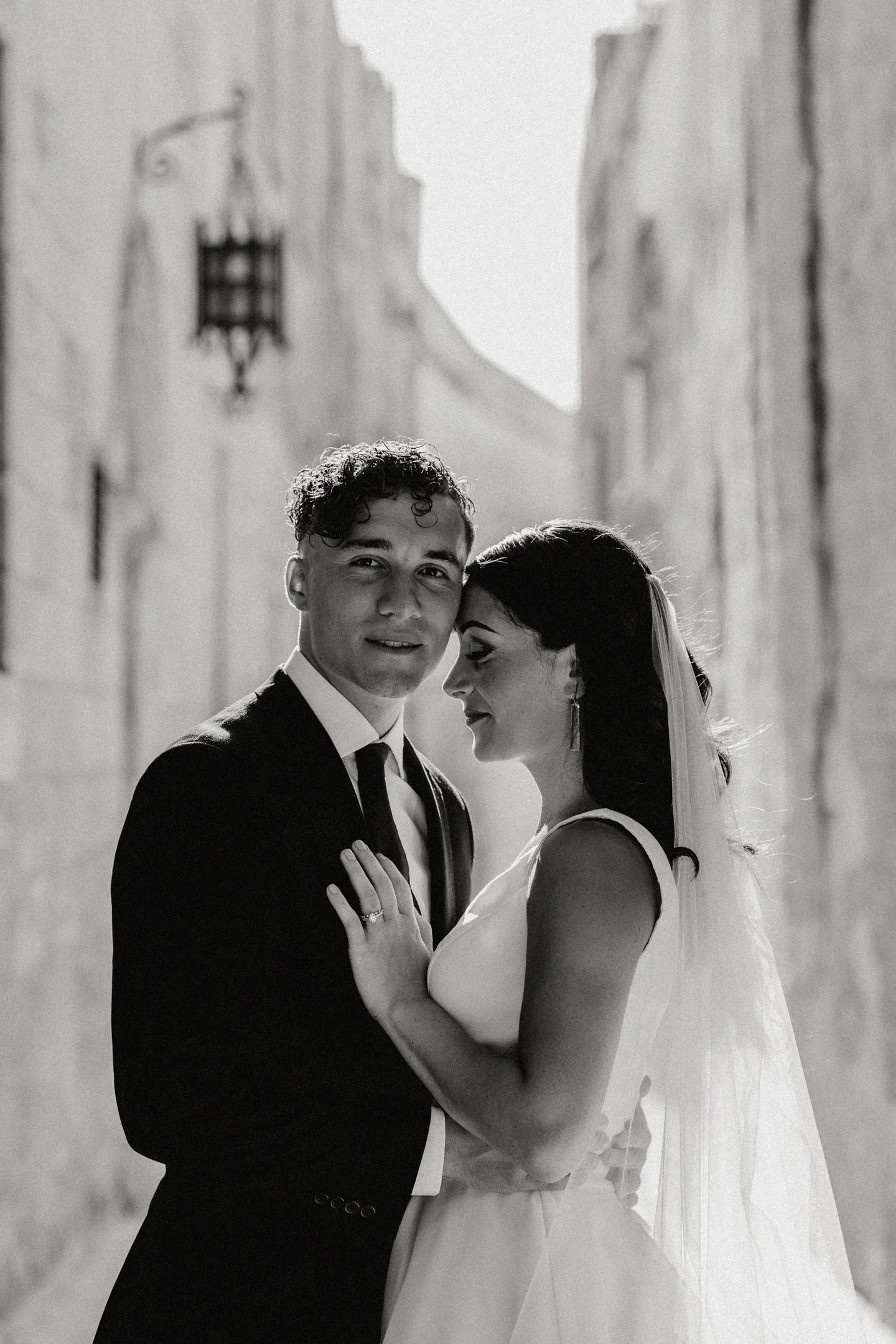 Black and white photograph of a newlywed couple standing close together in an alleyway, with the groom in a dark suit and the bride in a white wedding dress and veil, sharing an intimate moment in Malta, photographed by Dominique Paulette Photography