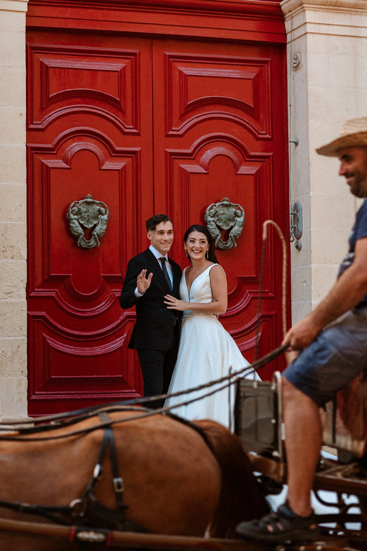 A newlywed couple stands in front of large, ornate red doors, smiling and waving, while a man on a horse-drawn carriage passes by in the foreground at their wedding in Malta, photographed by Dominique Paulette Photography.
