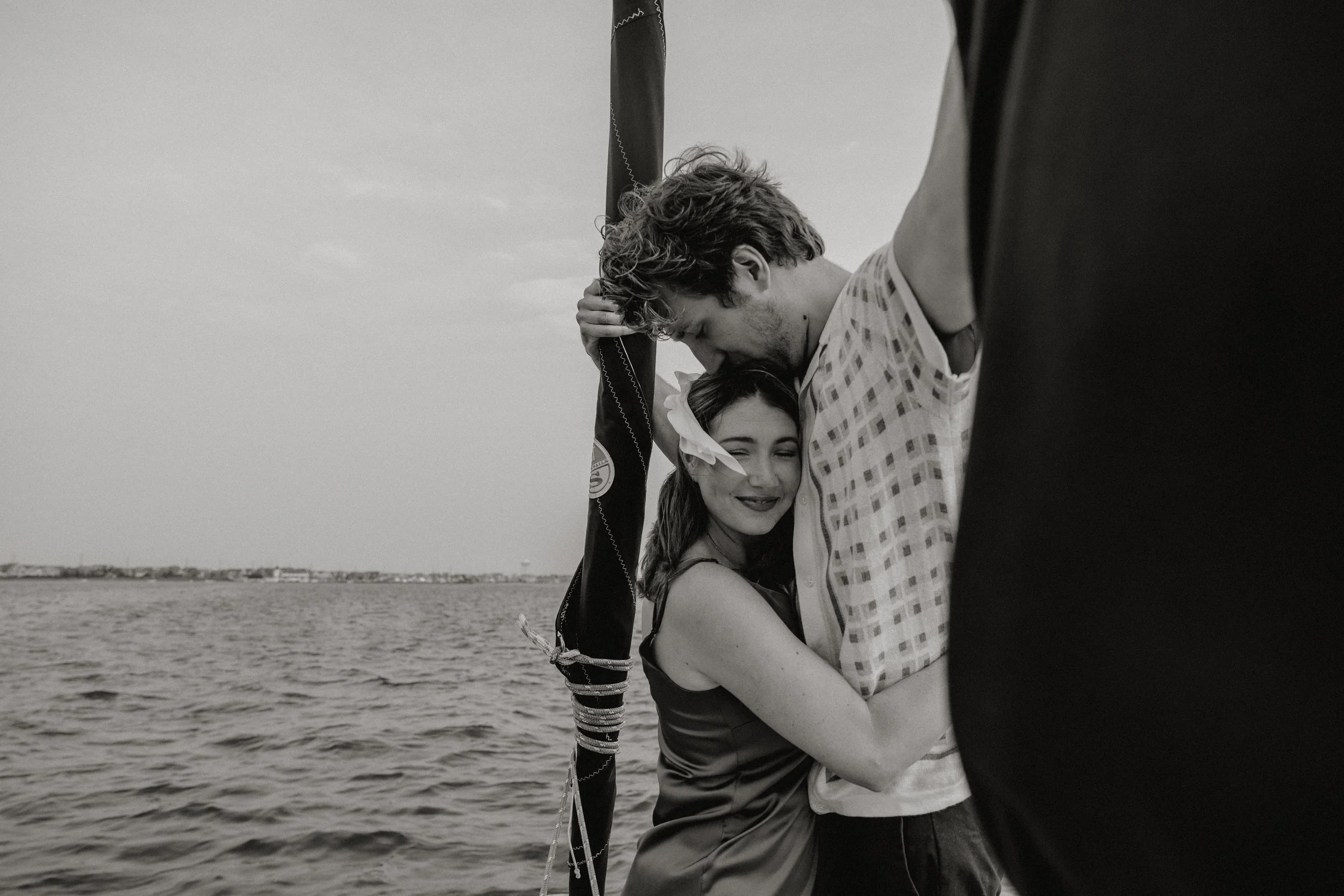 Black and white photo of a young couple embracing on a boat or dock, with water and a distant shoreline in the background, photographed by Dominique Paulette Photography