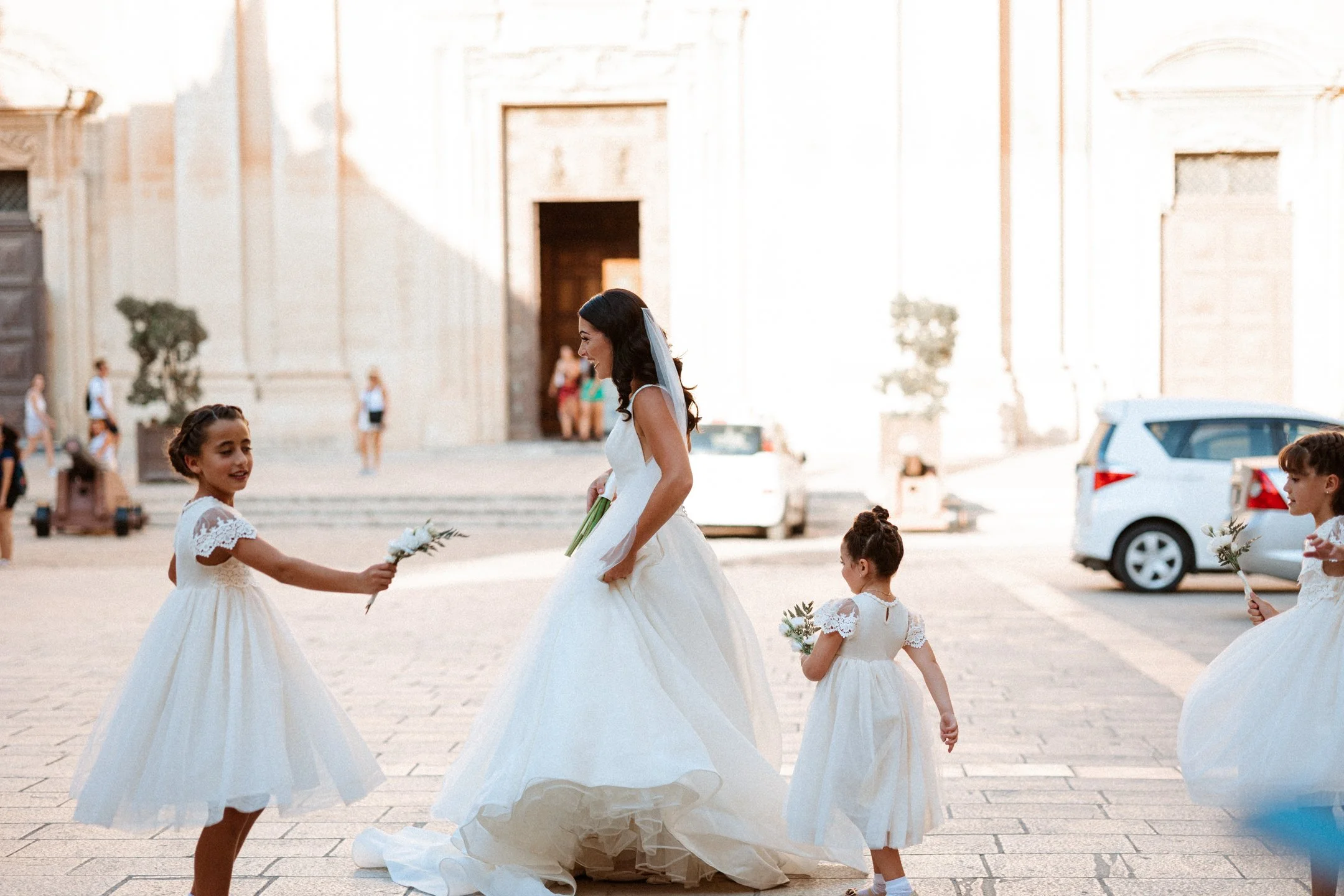 A bride in a white wedding gown holding a bouquet, surrounded by young girls in white dresses holding flowers, outside in front of a large historic building for their wedding in Malta, photographed by Dominique Paulette Photography