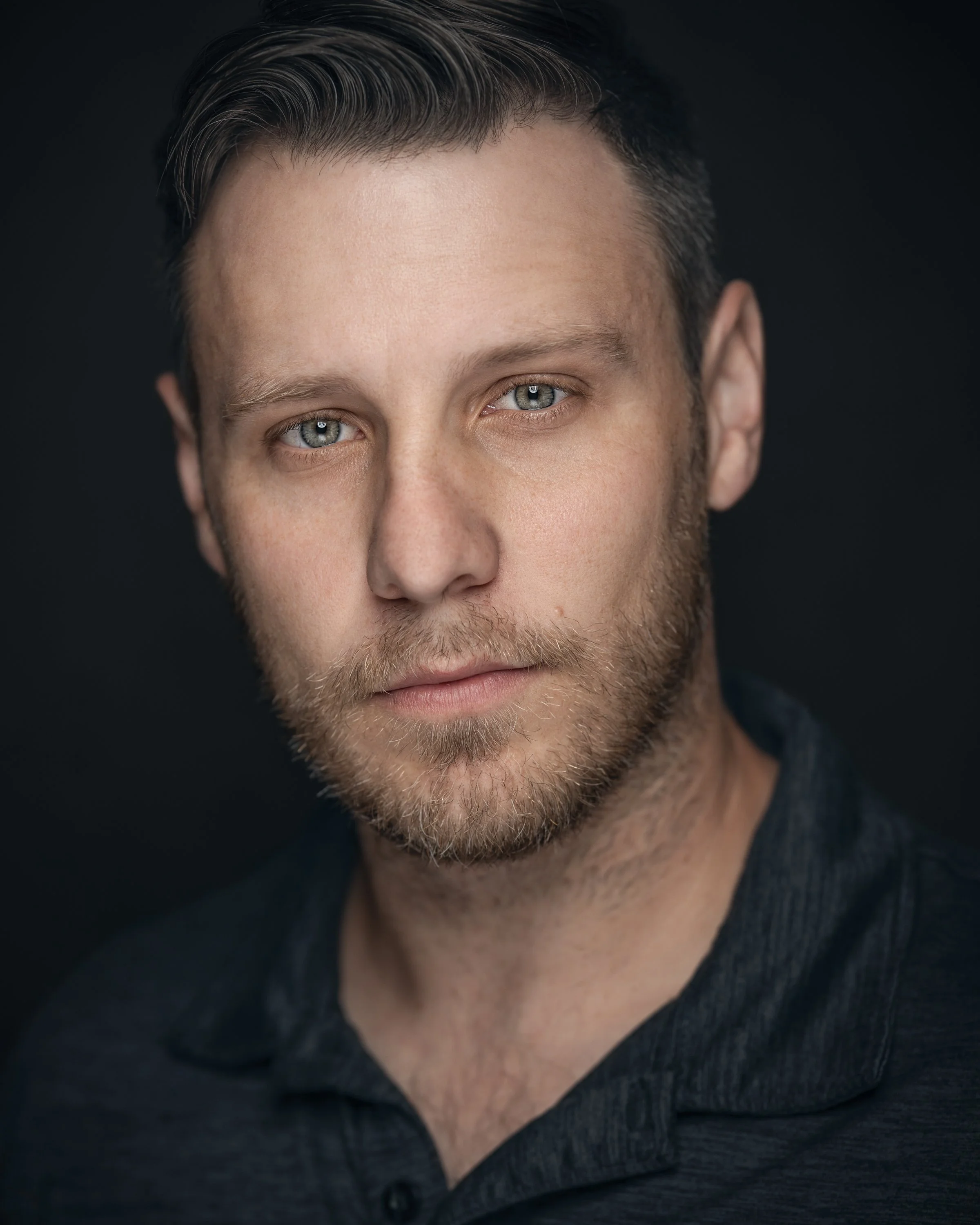 Close-up portrait of a young man with light hair and beard, wearing a dark shirt, against a dark background.