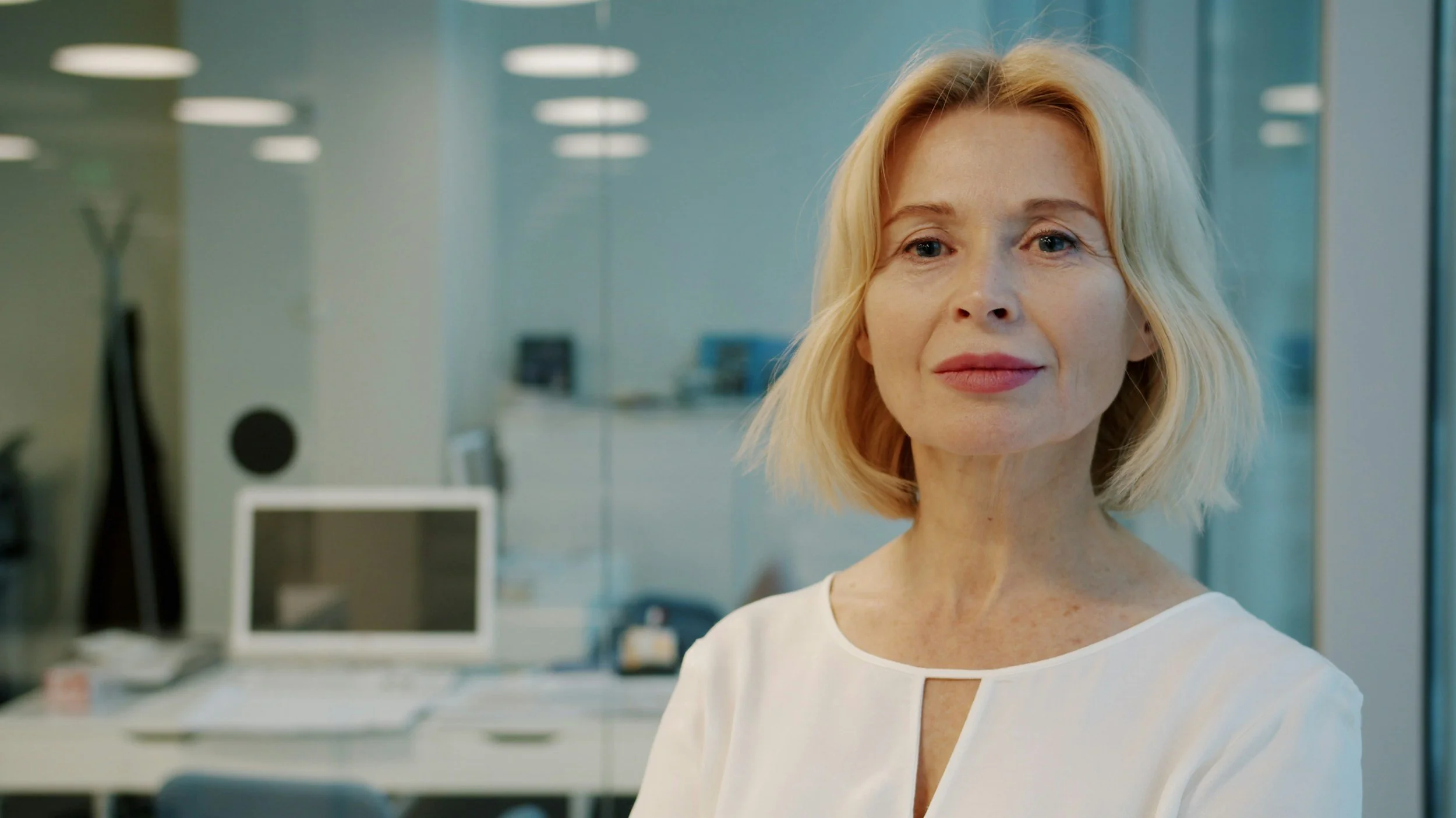 A woman with blonde hair and a white shirt standing in an office, looking directly at the camera.