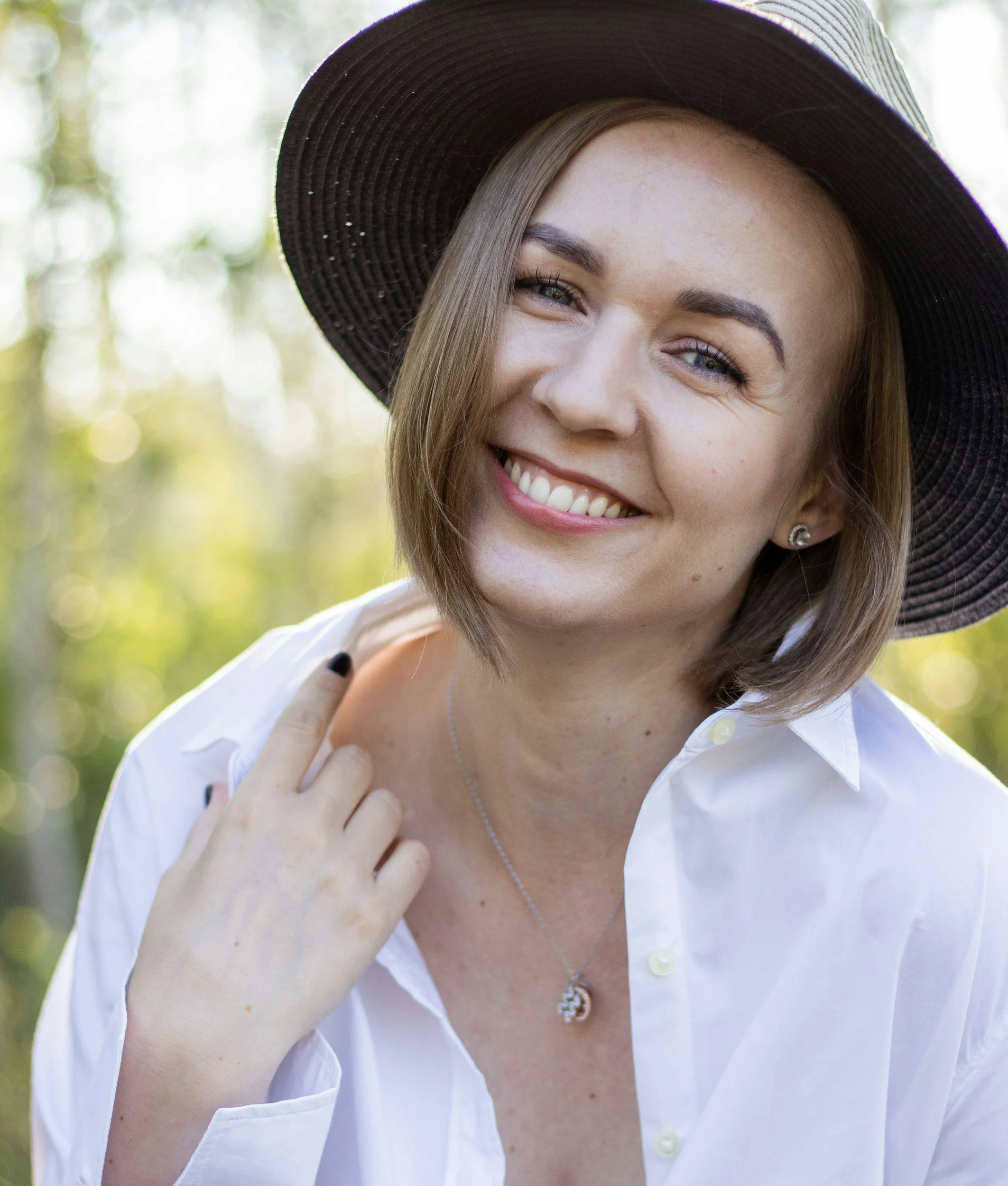 A smiling woman with short brown hair wearing a large black sun hat, a white button-up shirt, a necklace, and earrings, outdoors in a sunlit natural setting.