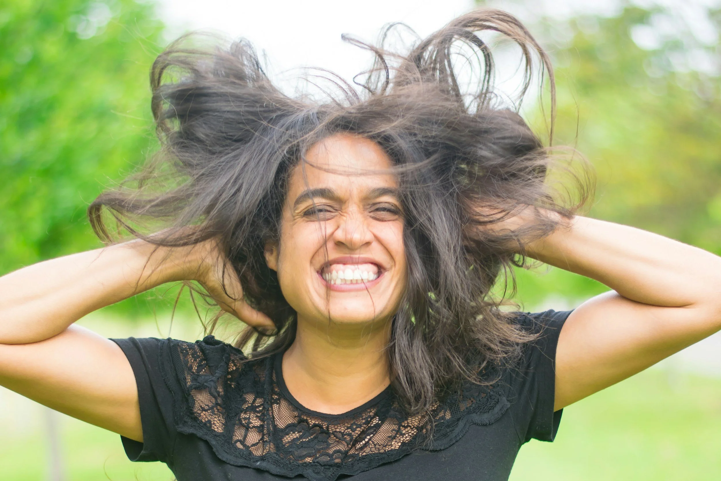 A woman smiling with her eyes closed and hair flying outdoors in a park.