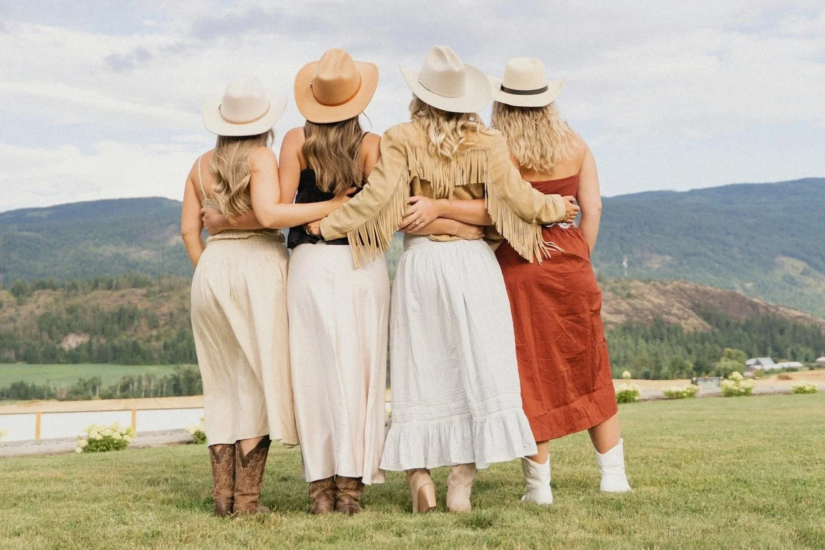 Five women in cowboy hats and western attire standing together with their arms around each other outdoors, with mountains and a cloudy sky in the background.