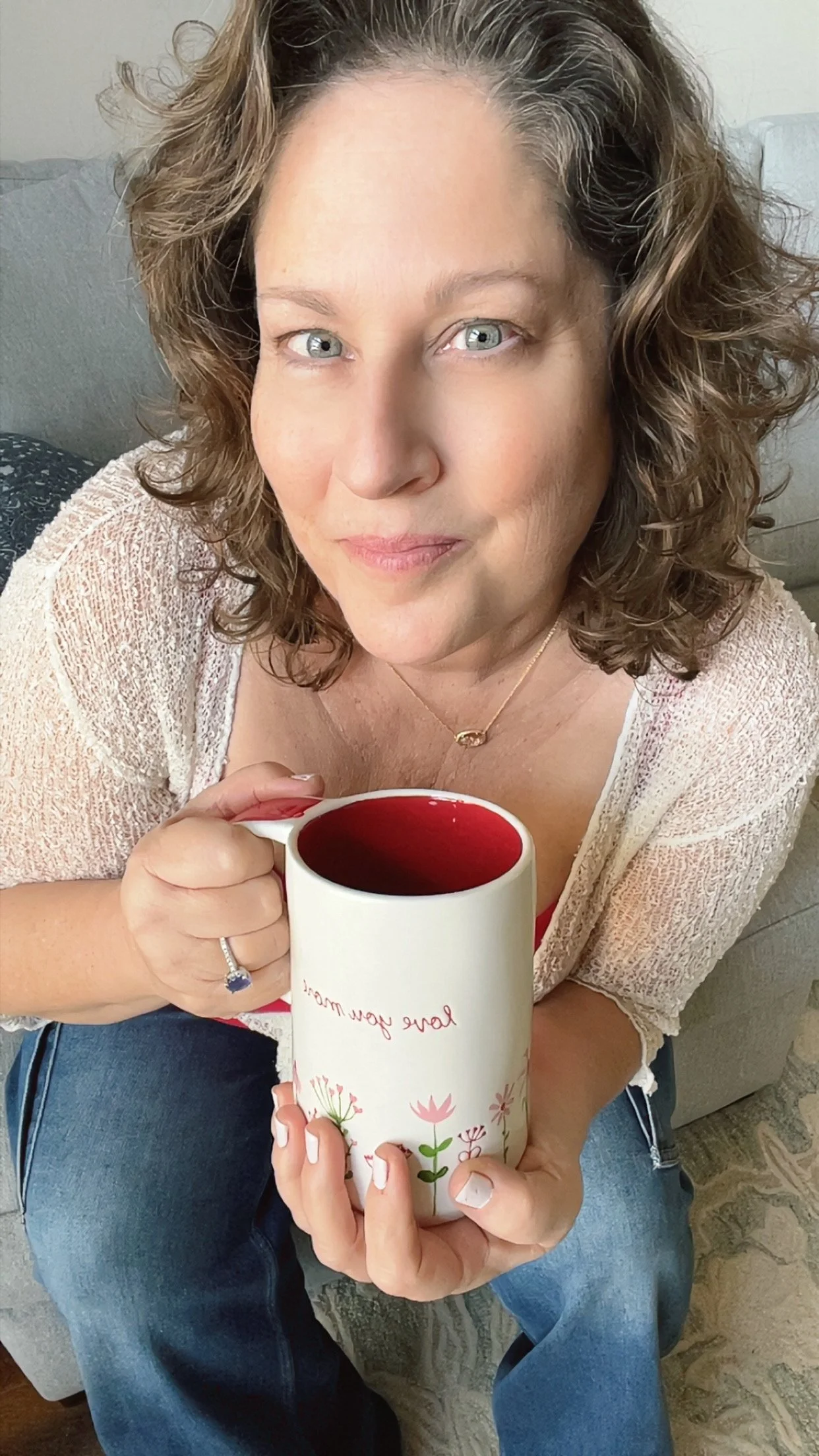 A woman with curly hair and blue eyes holding a white mug with red inside, sitting on a couch.