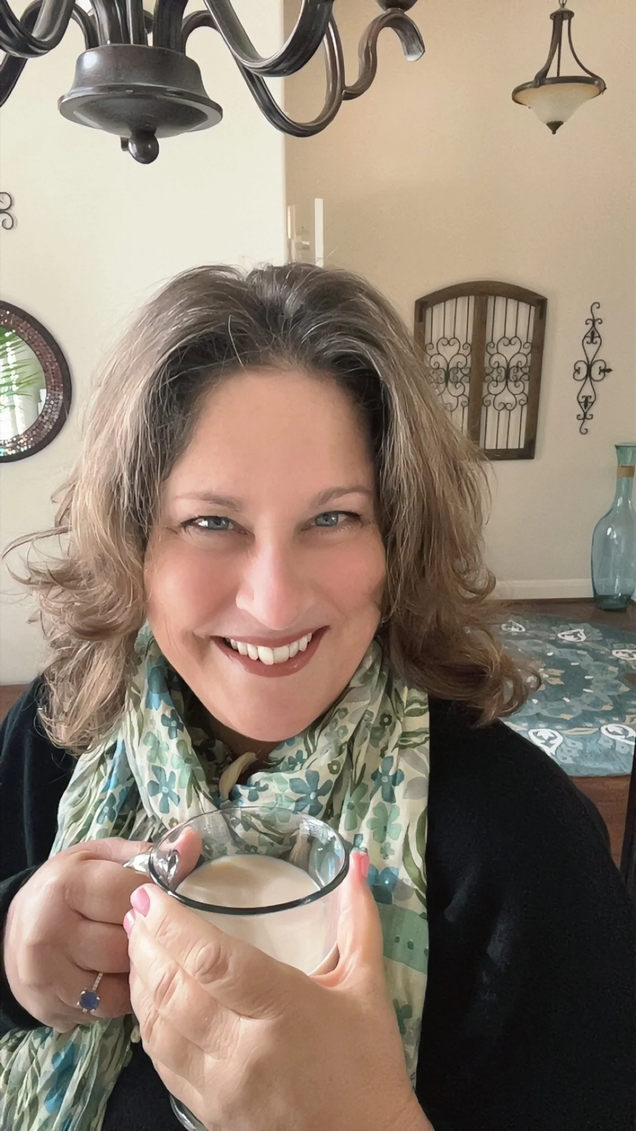 A smiling woman with wavy hair holding a glass of coffee or tea, sitting in a dining room with a chandelier, decorative wall hangings, and a table with a patterned tablecloth.