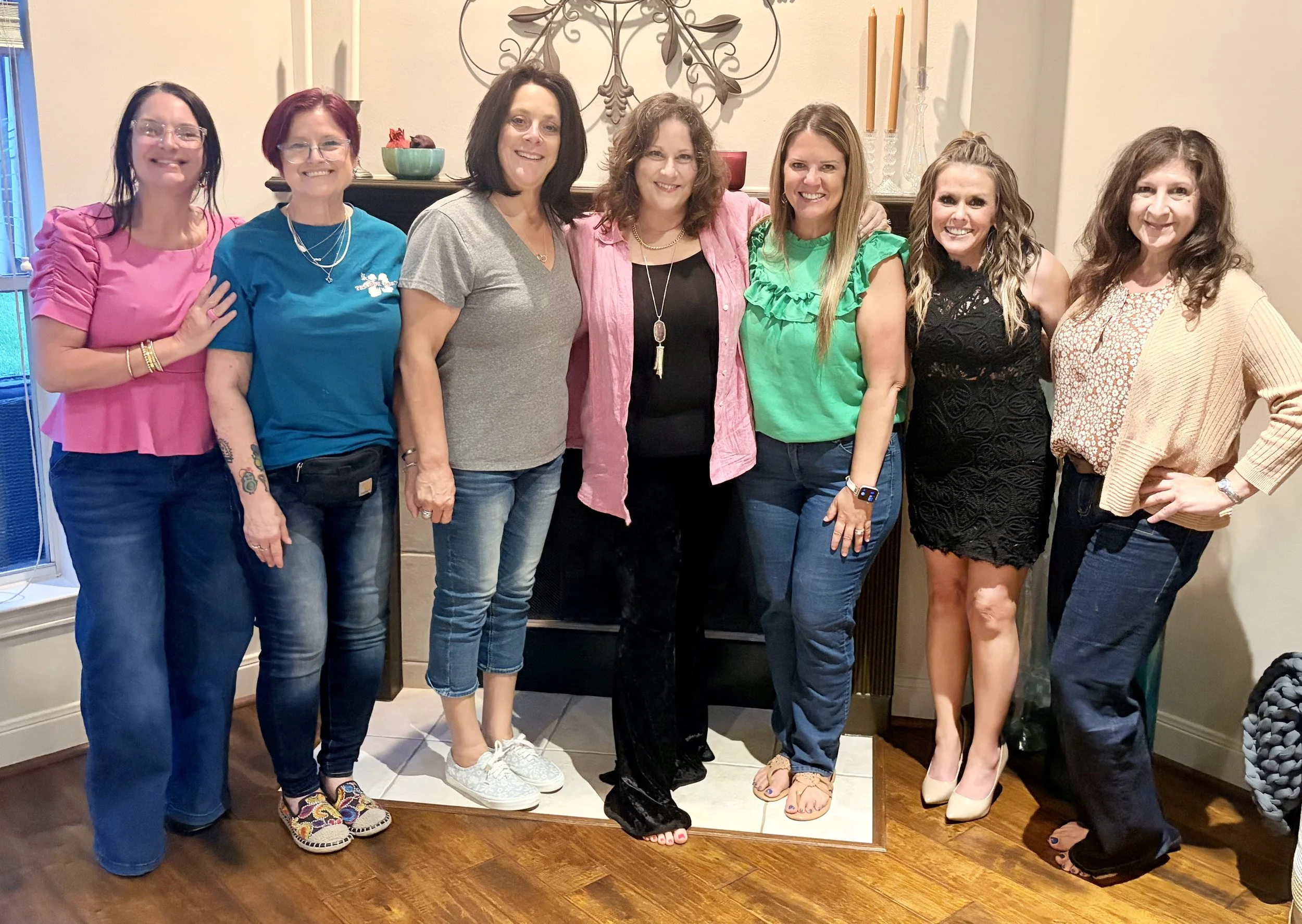 A group of eight women standing together in a living room, posing for a photo. They are smiling and dressed casually or semi-formally. The background features a decorative wall piece, candles, and a fireplace.