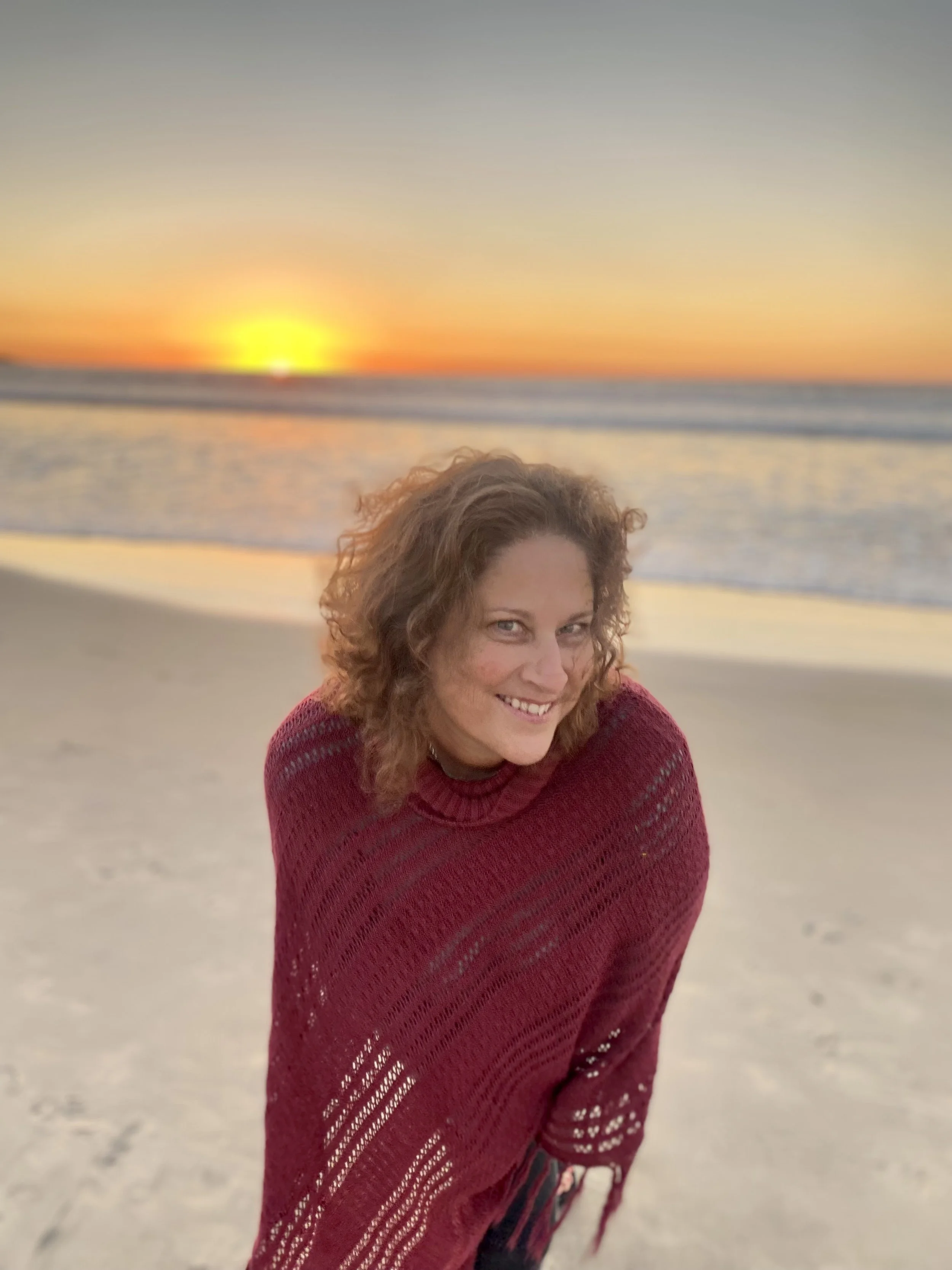 A lady with brown hair standing on the beach with a sunrise behind her.