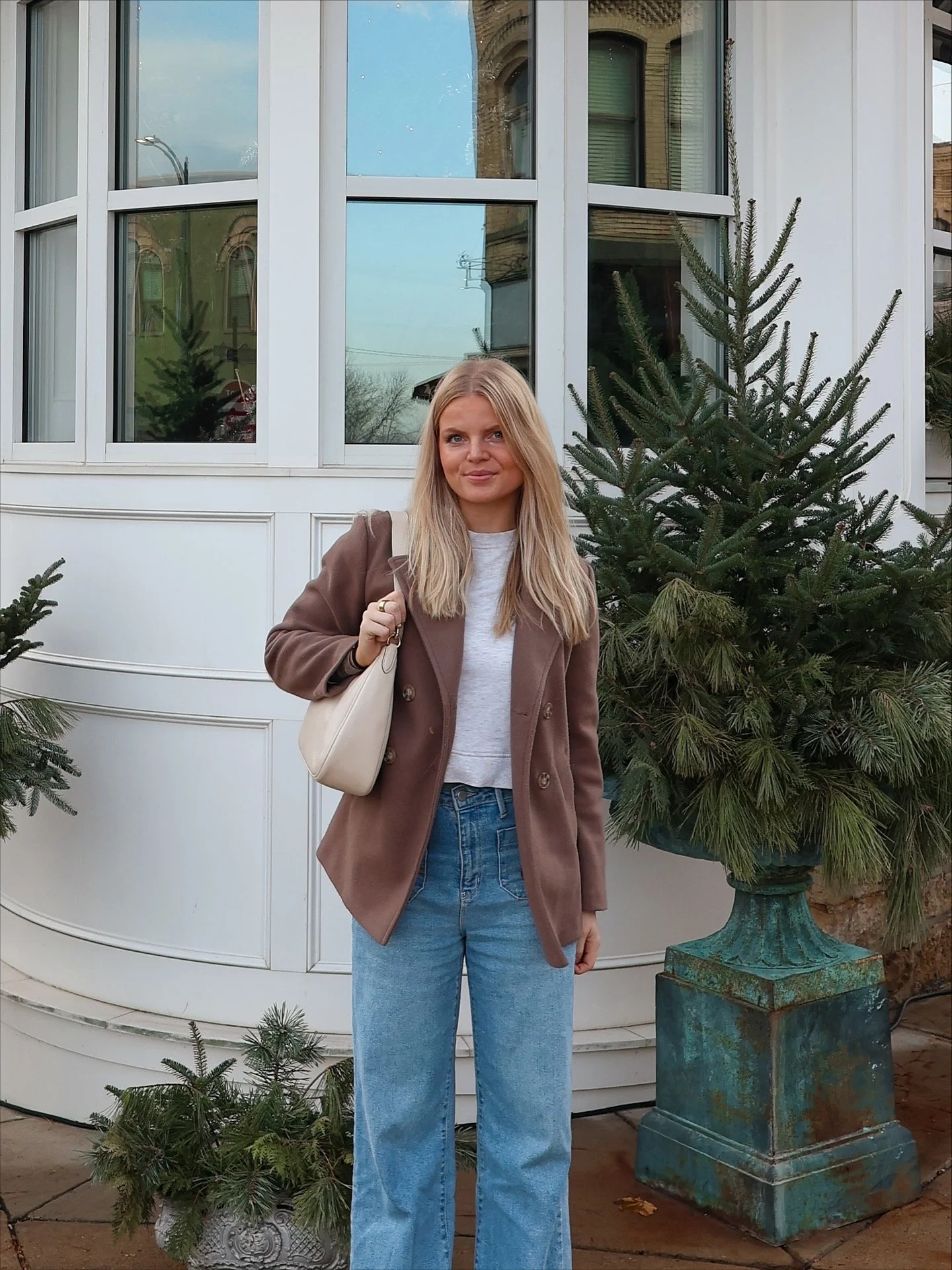 A young woman with long blonde hair, wearing a brown coat, grey sweatshirt, and blue jeans, standing outdoors in front of a white building with large windows, surrounded by Christmas trees.