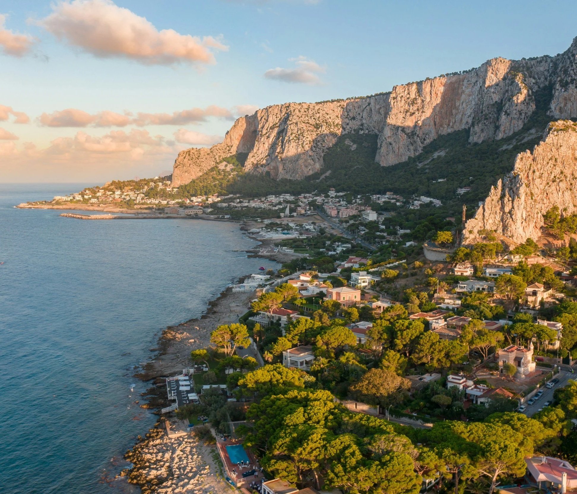 Aerial view of a coastal town with green trees, white buildings, and rocky cliffs at sunset.