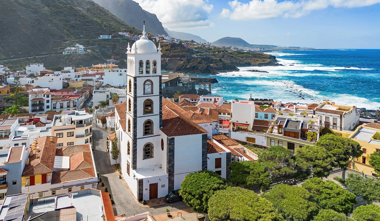 An aerial view of a coastal town with white buildings and orange-tiled roofs, centered around a white church with a tall bell tower and a clock, overlooking the ocean with waves and rocky shoreline, with green trees and mountainous terrain in the bac