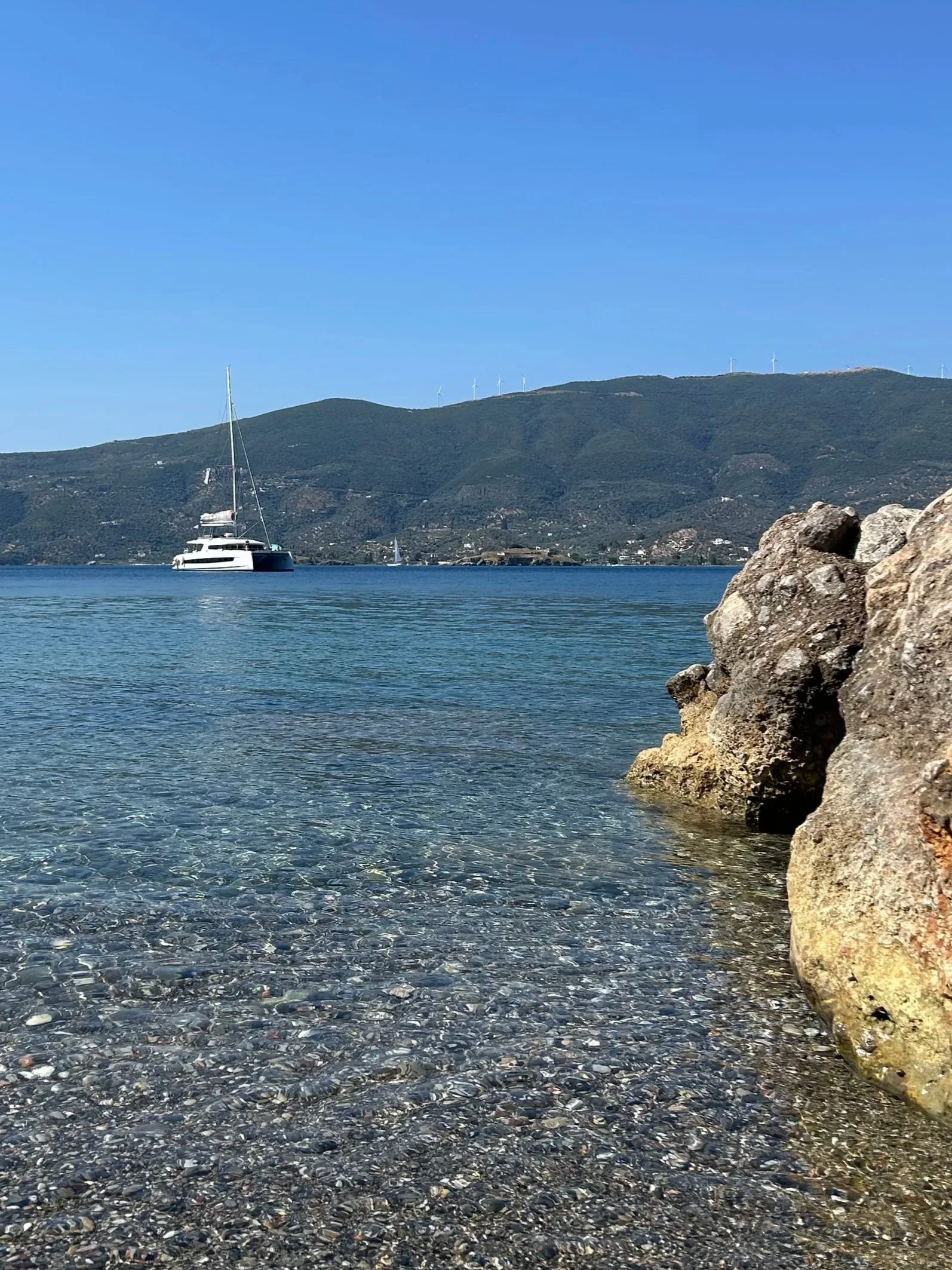 Calm water with a large white yacht, green hills in the background with wind turbines, and rocks in the foreground.